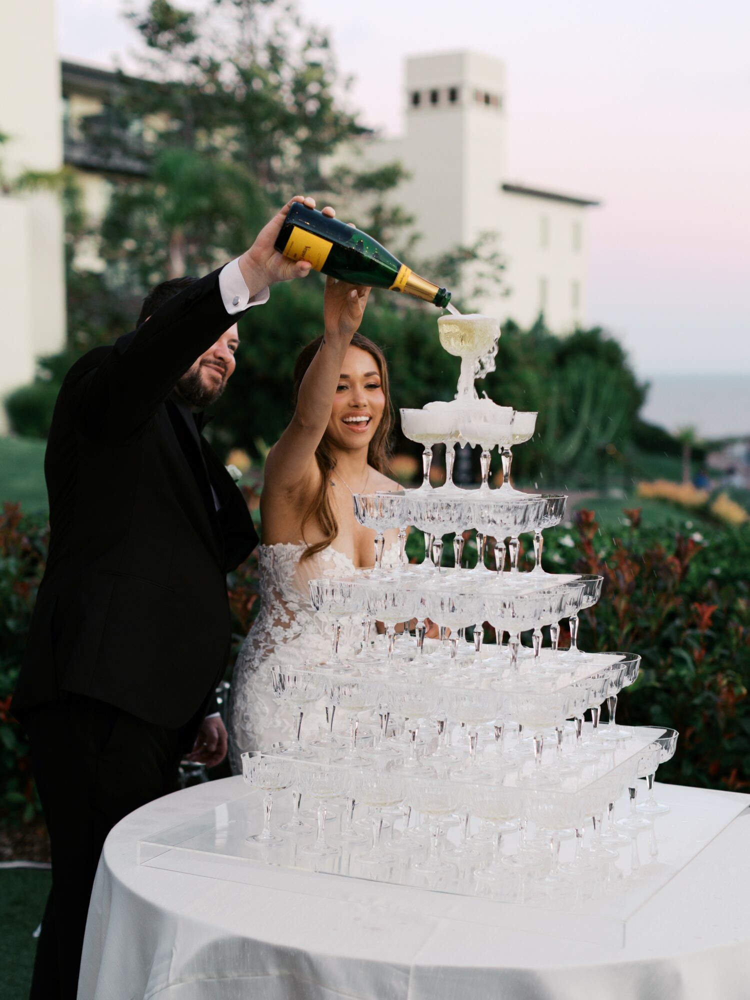 A man and woman in formal attire pour champagne into a multi-tiered ice sculpture cake outdoors.