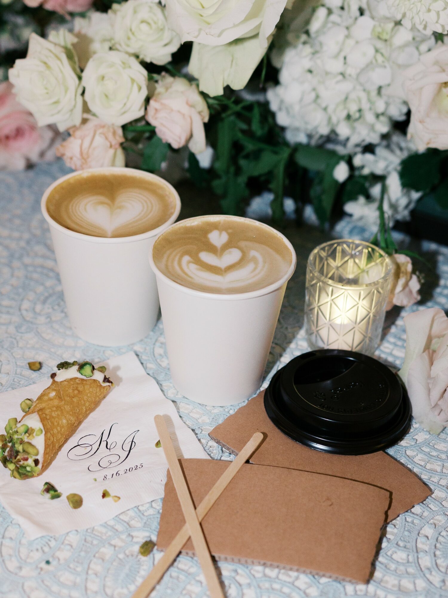 Two cups of coffee with heart-shaped latte art, surrounded by flowers, a candle, and a black lid on a table.