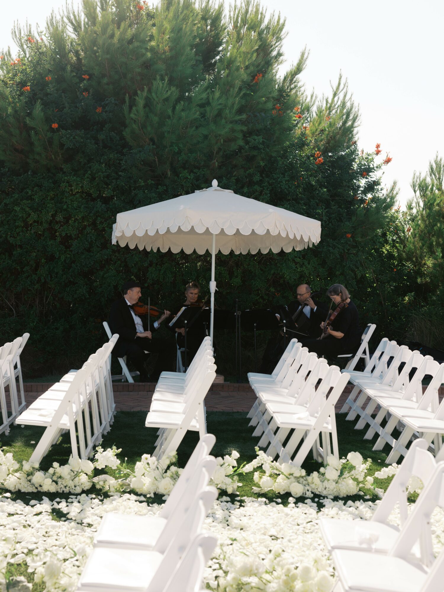White chairs arranged in rows with floral decorations, umbrella in the center, people standing and talking in the background.