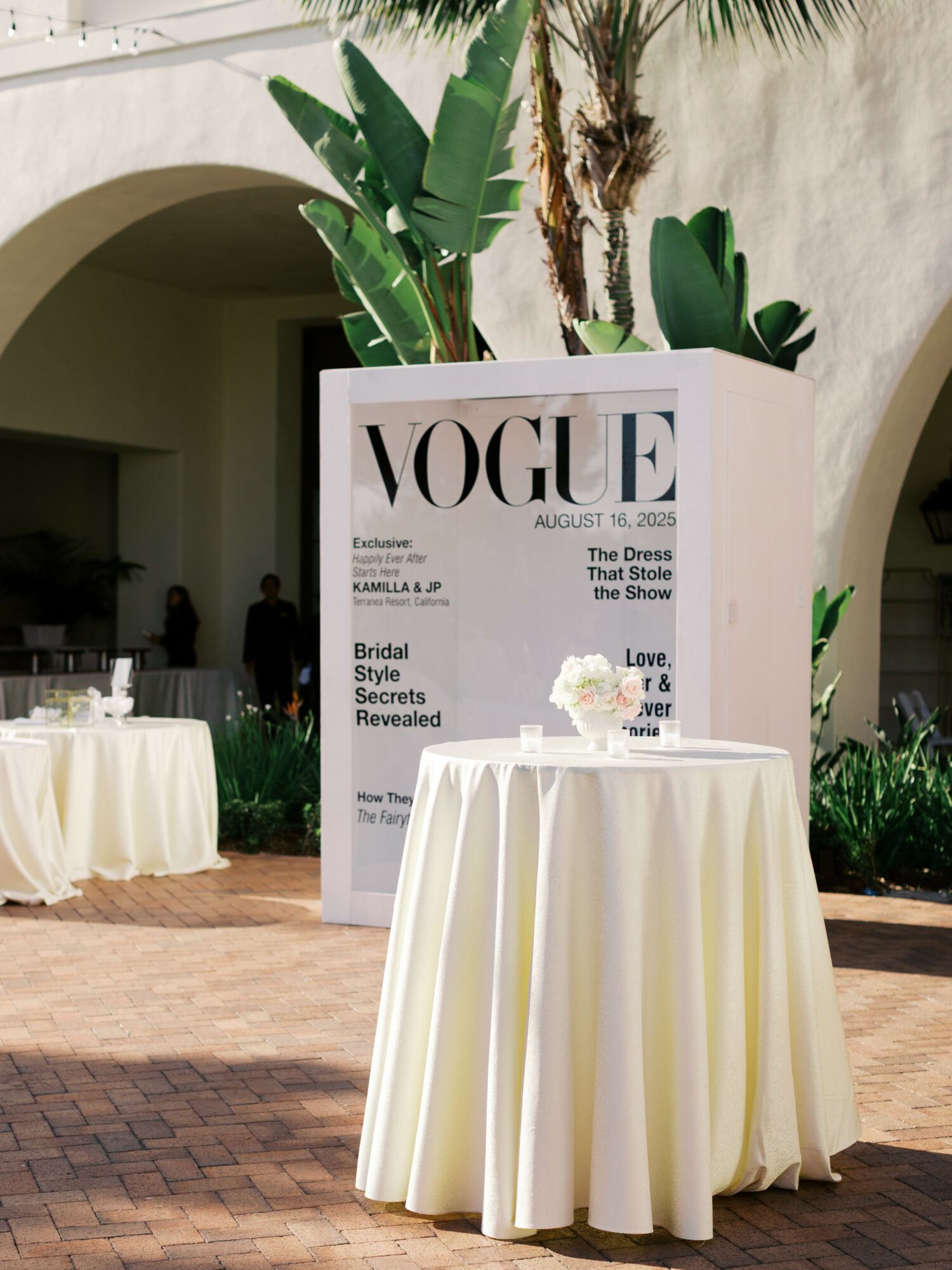 Round table with white tablecloth and floral centerpiece outdoors, large Vogue magazine cover sign in background, greenery and white building.