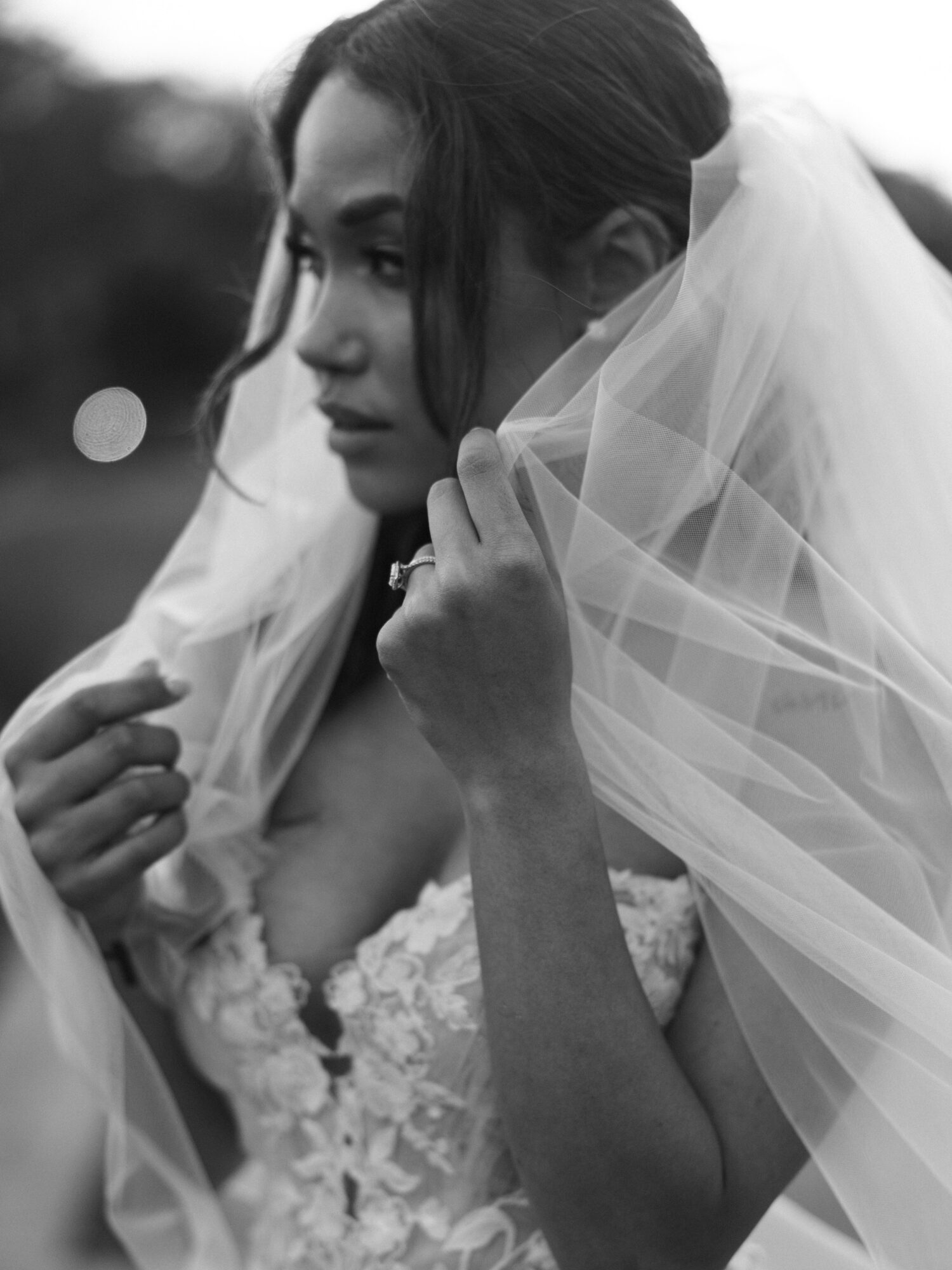 Woman in wedding dress holding veil, looking to the side outdoors, black and white photo.