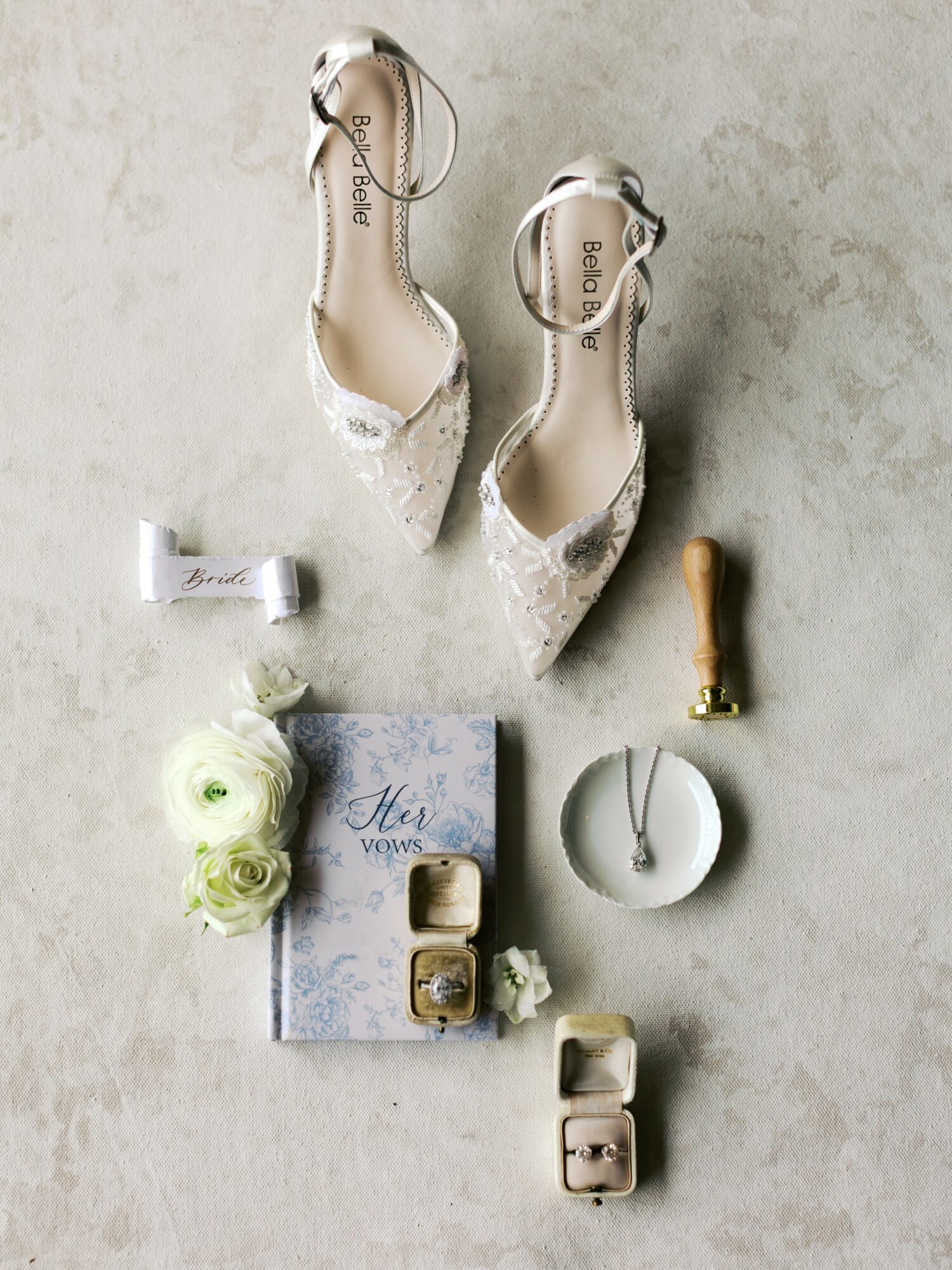 Pair of white lace high heels, jewelry, flowers, and a book on a light surface.