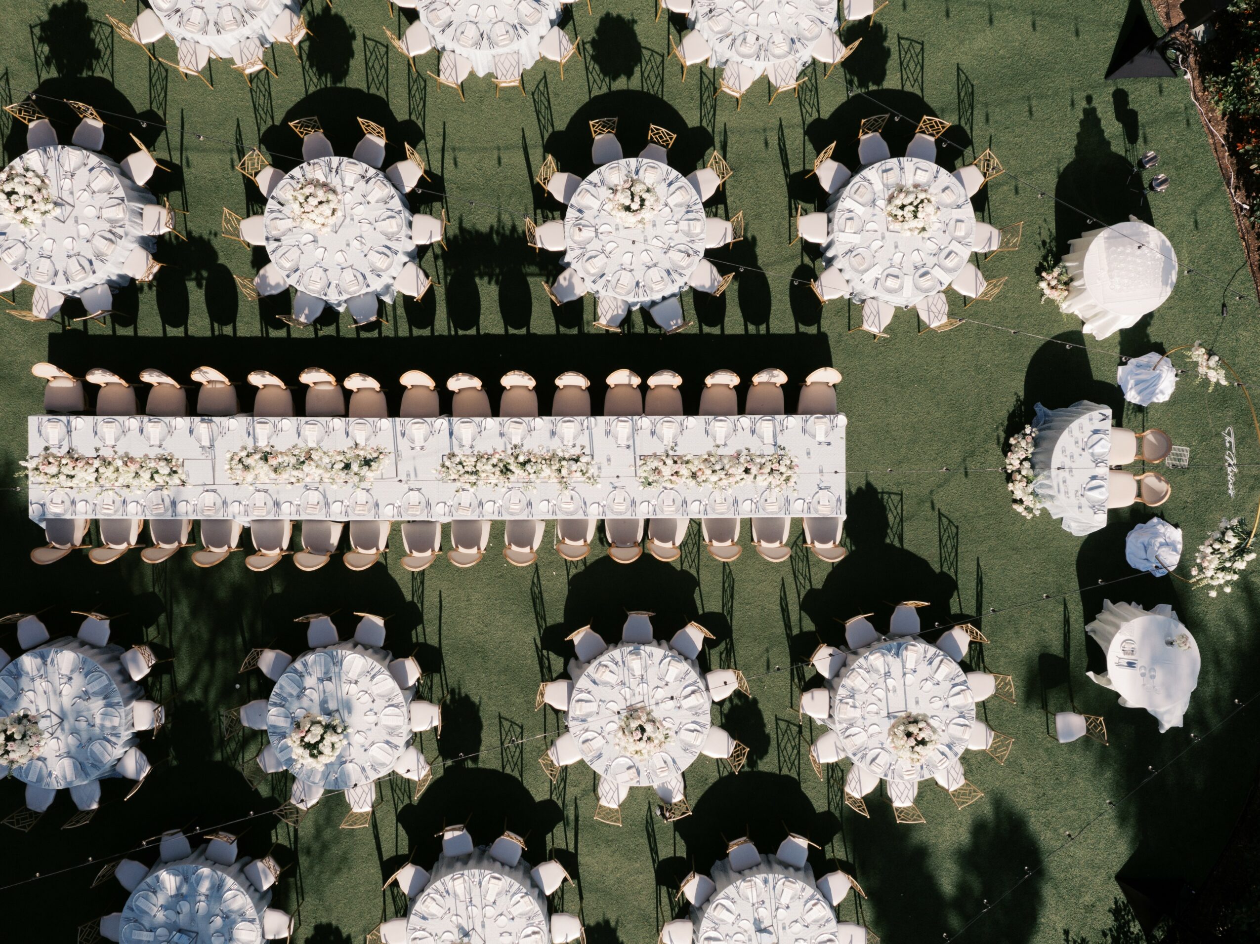 Outdoor event setup with round tables, chairs, and umbrellas on grass, some tables with centerpieces.