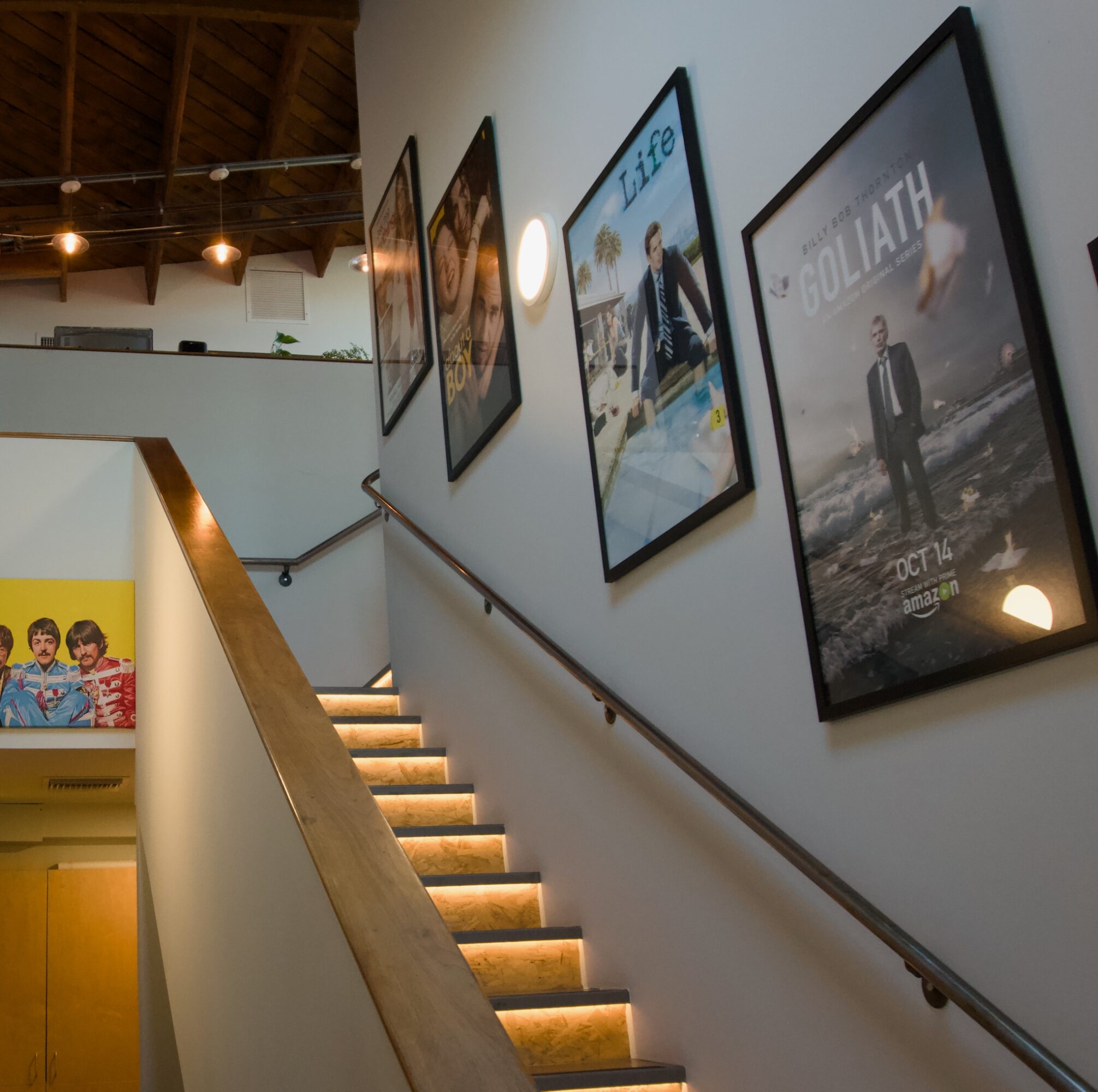 Staircase with illuminated steps and framed posters on the wall, viewed from below.