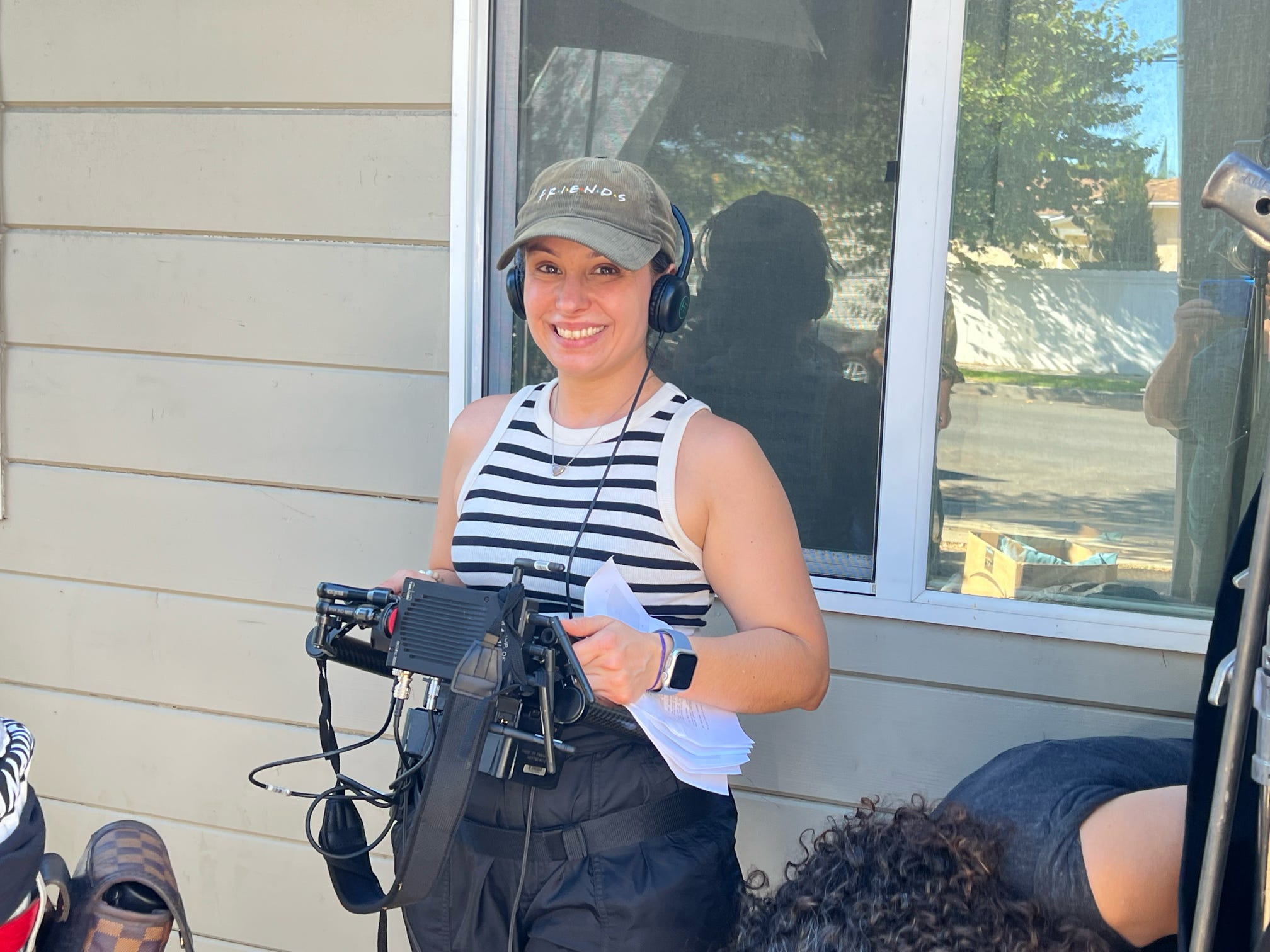 Young woman wearing a striped shirt, cap, and headphones, smiling outdoors near a window.