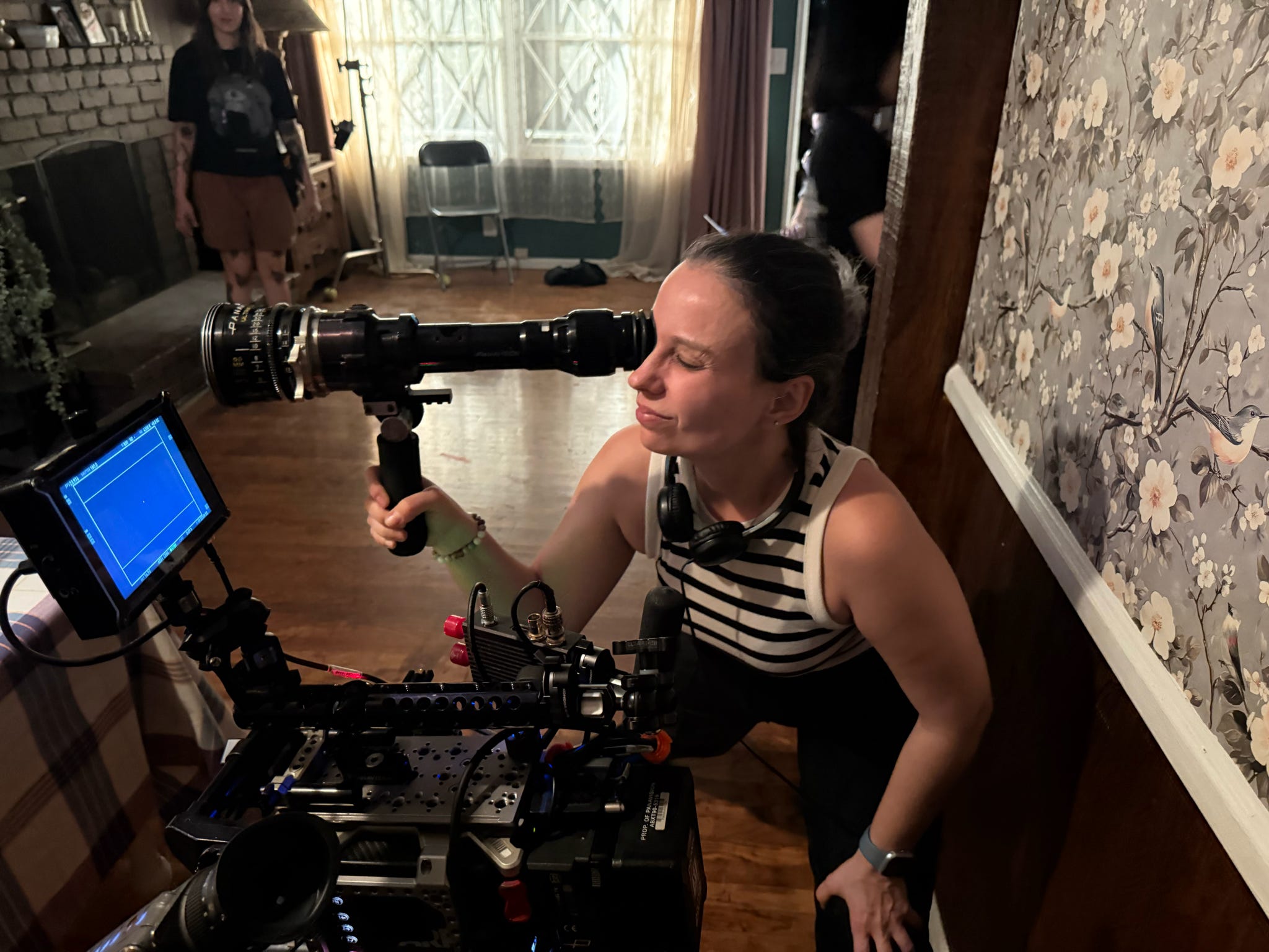 Woman operating camera equipment indoors, with people and furniture in background.