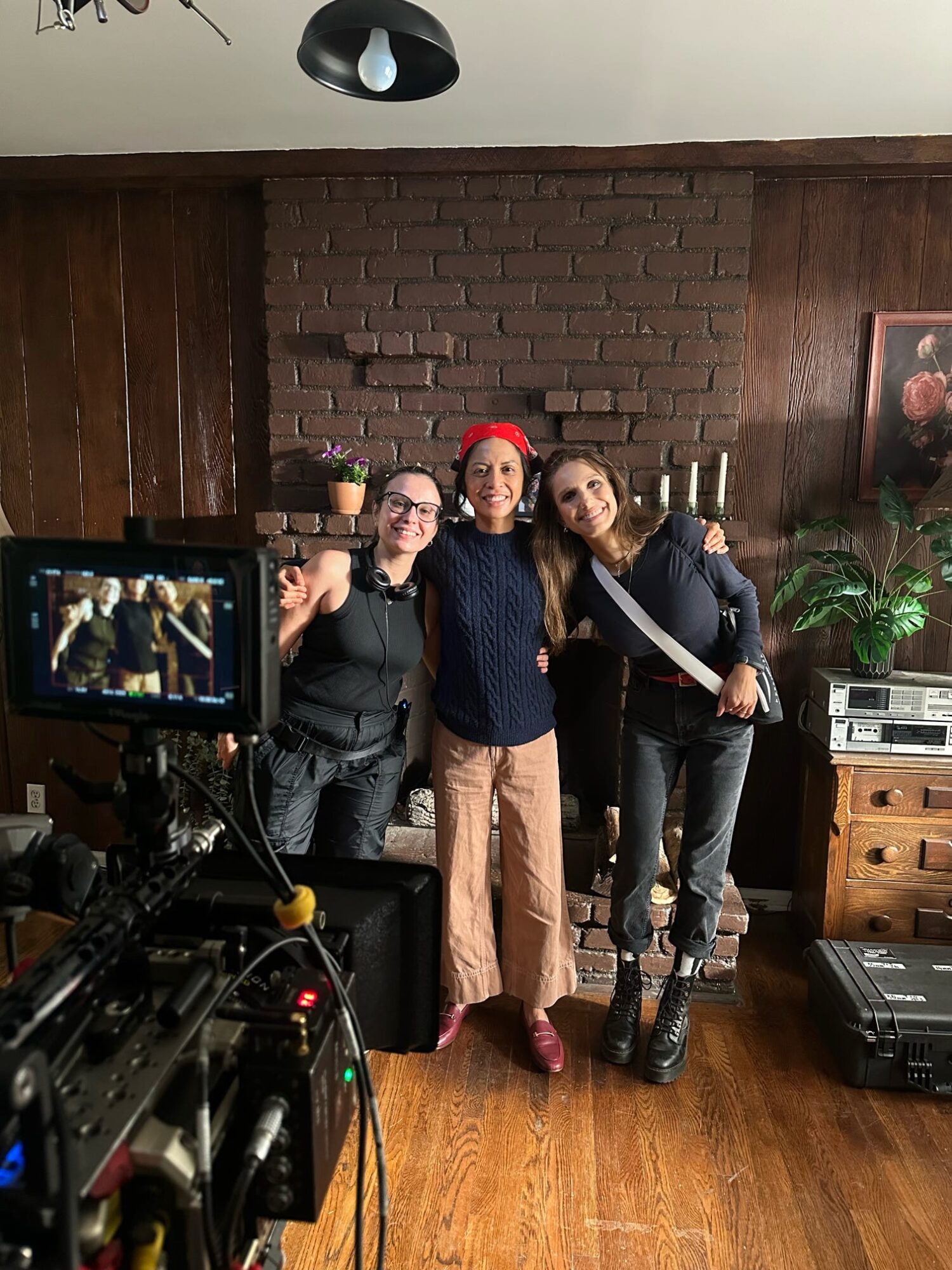 Three women standing together indoors, smiling, with a brick wall, wooden paneling, and a plant in the background.