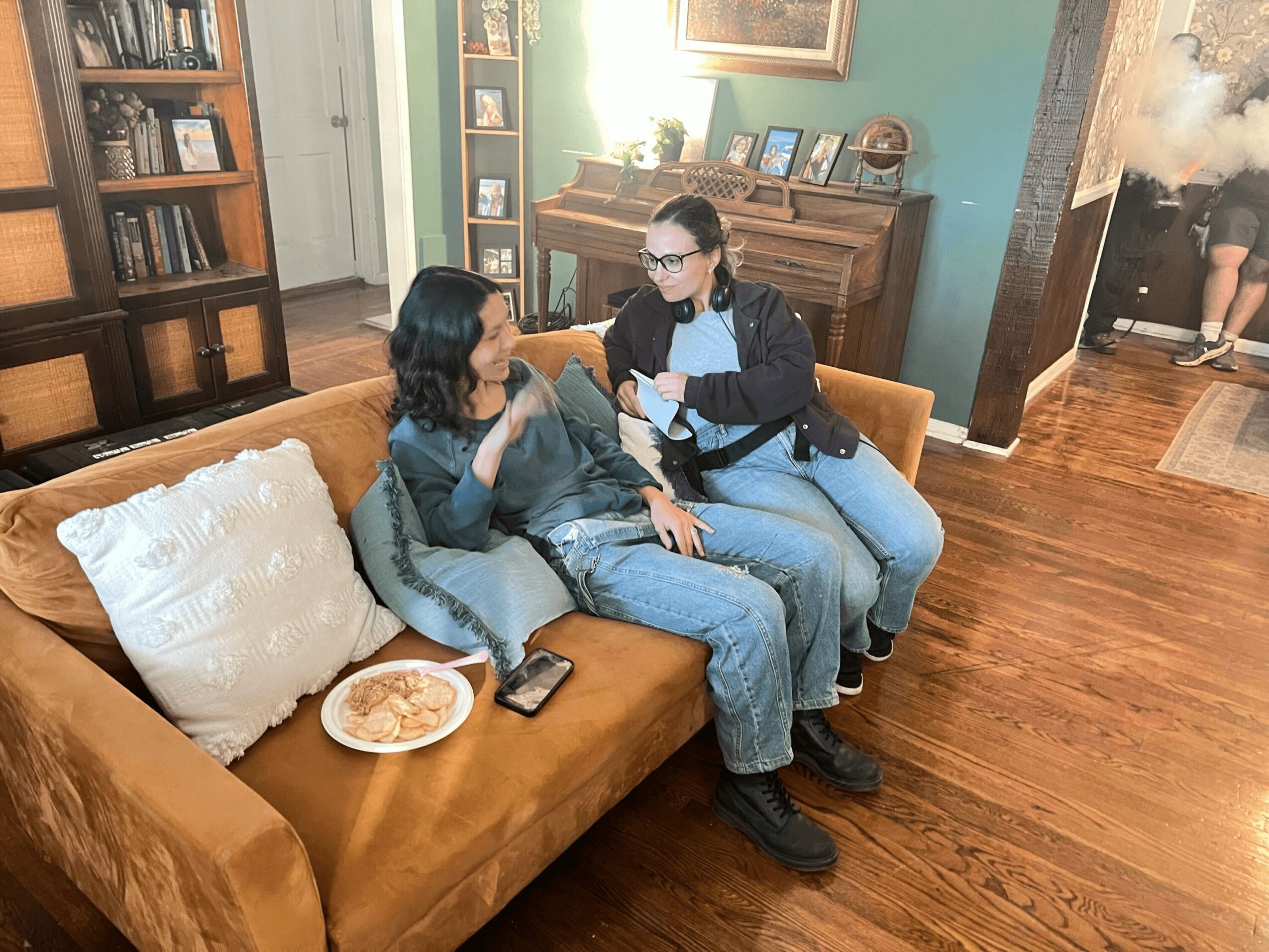 Two women sit on a brown couch talking, one holding a phone, in a cozy living room with wooden floors and bookshelves.
