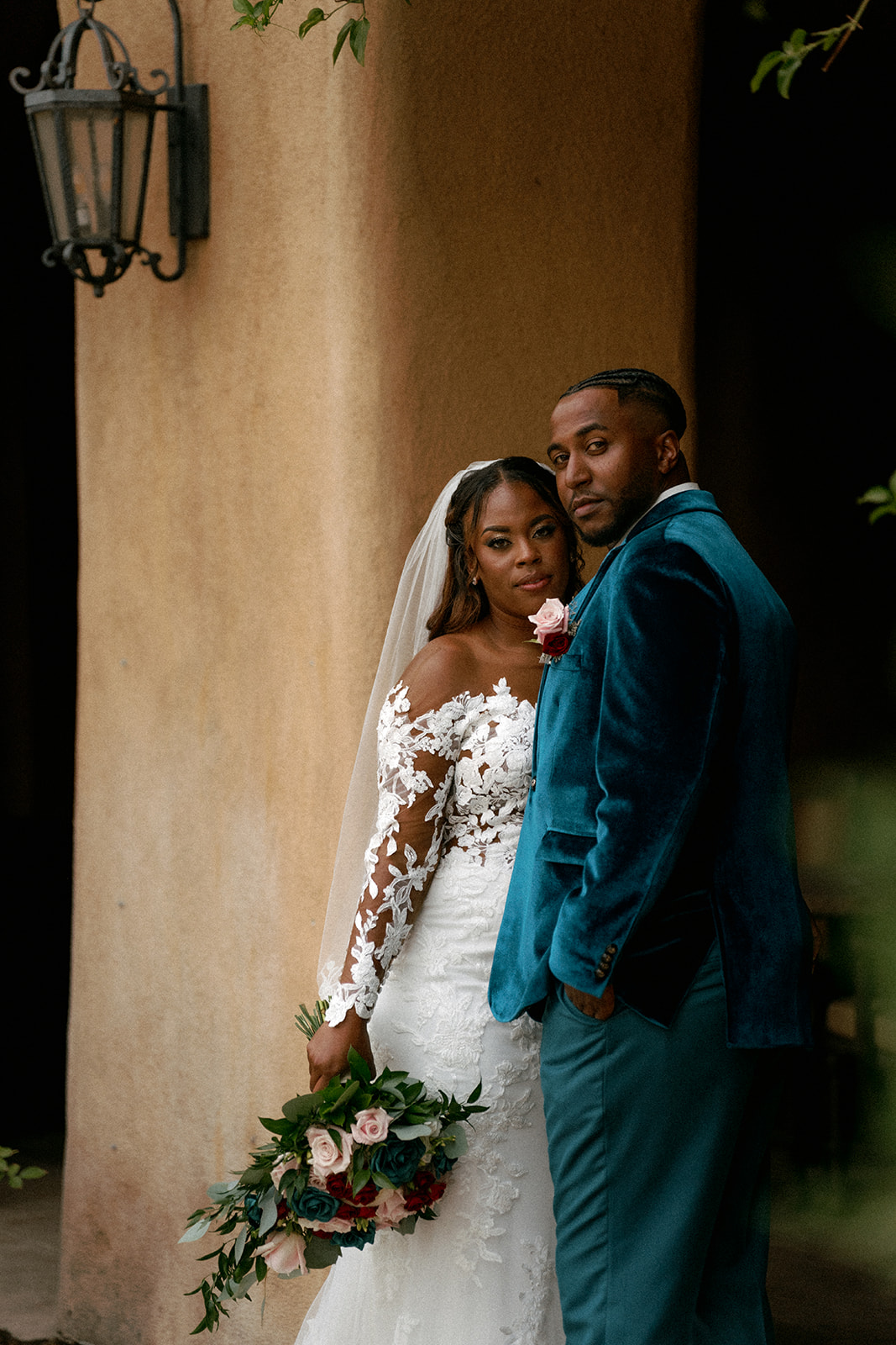Bride and groom stand close together outdoors, bride holding a bouquet, both looking at camera, with a wall and lantern in background.