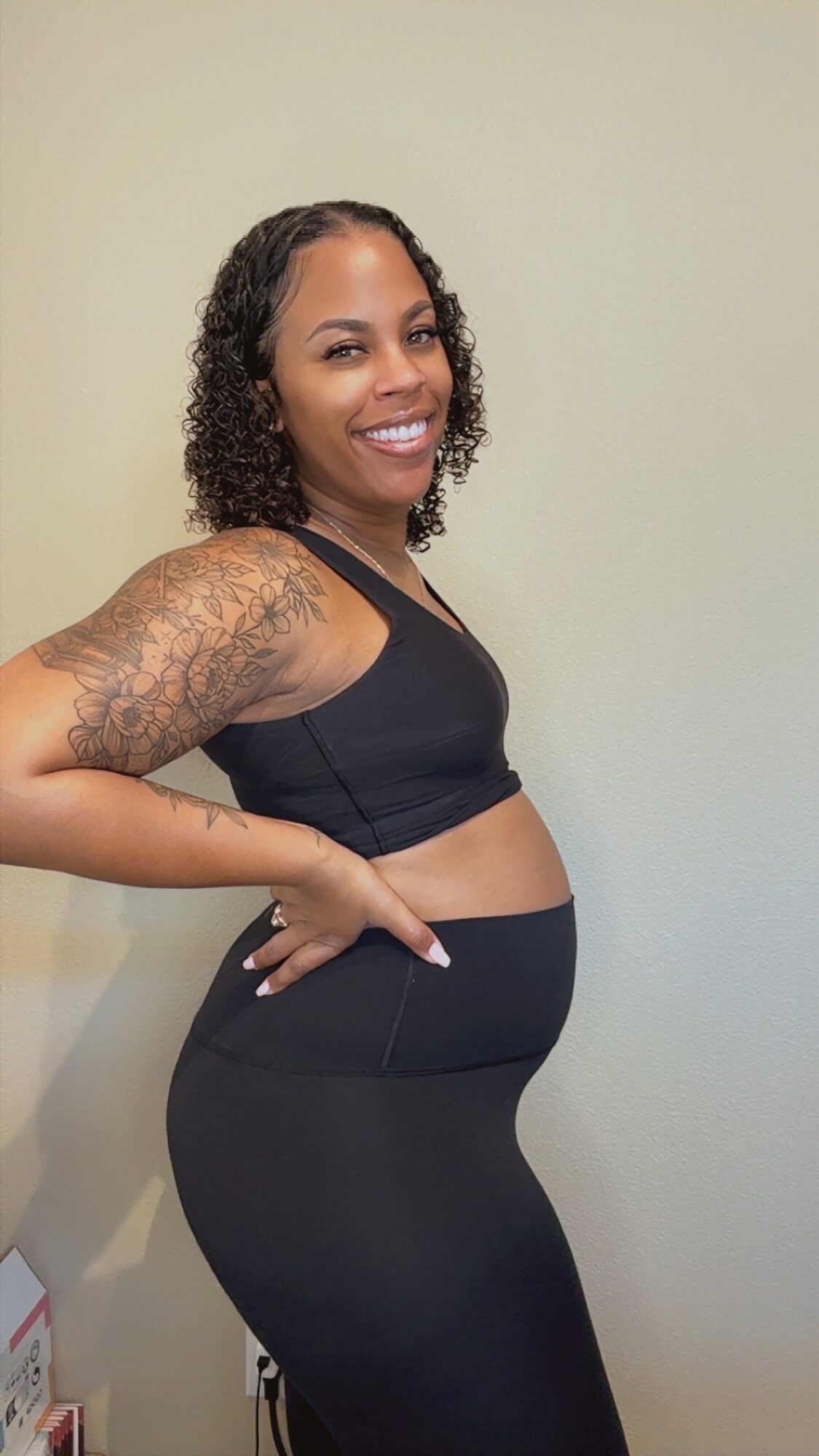Smiling woman with curly hair and tattoos on her arm, wearing black sports bra and leggings, standing sideways against a plain wall.