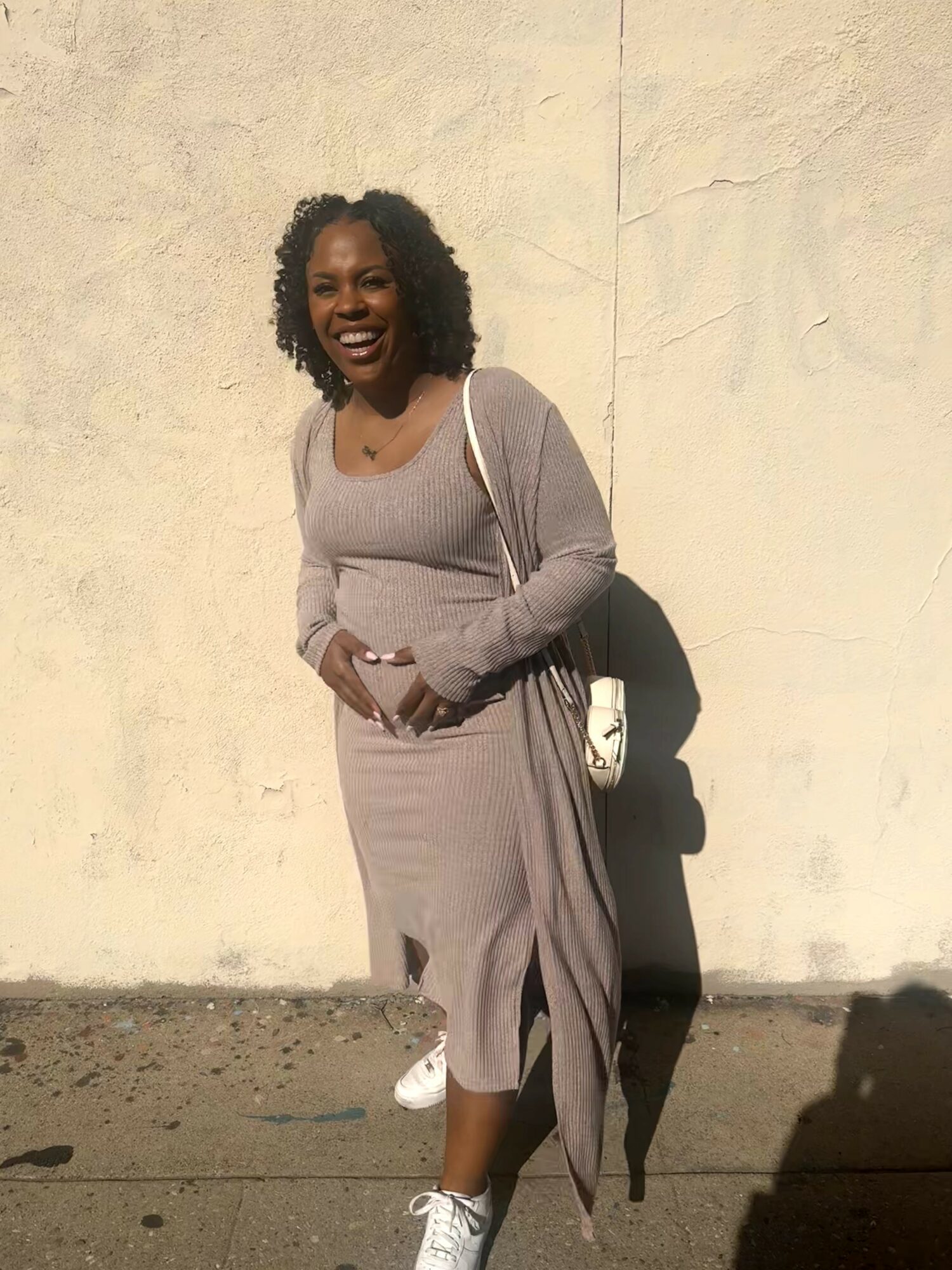 Woman smiling, standing against a light-colored wall, wearing a long dress and carrying a bag.