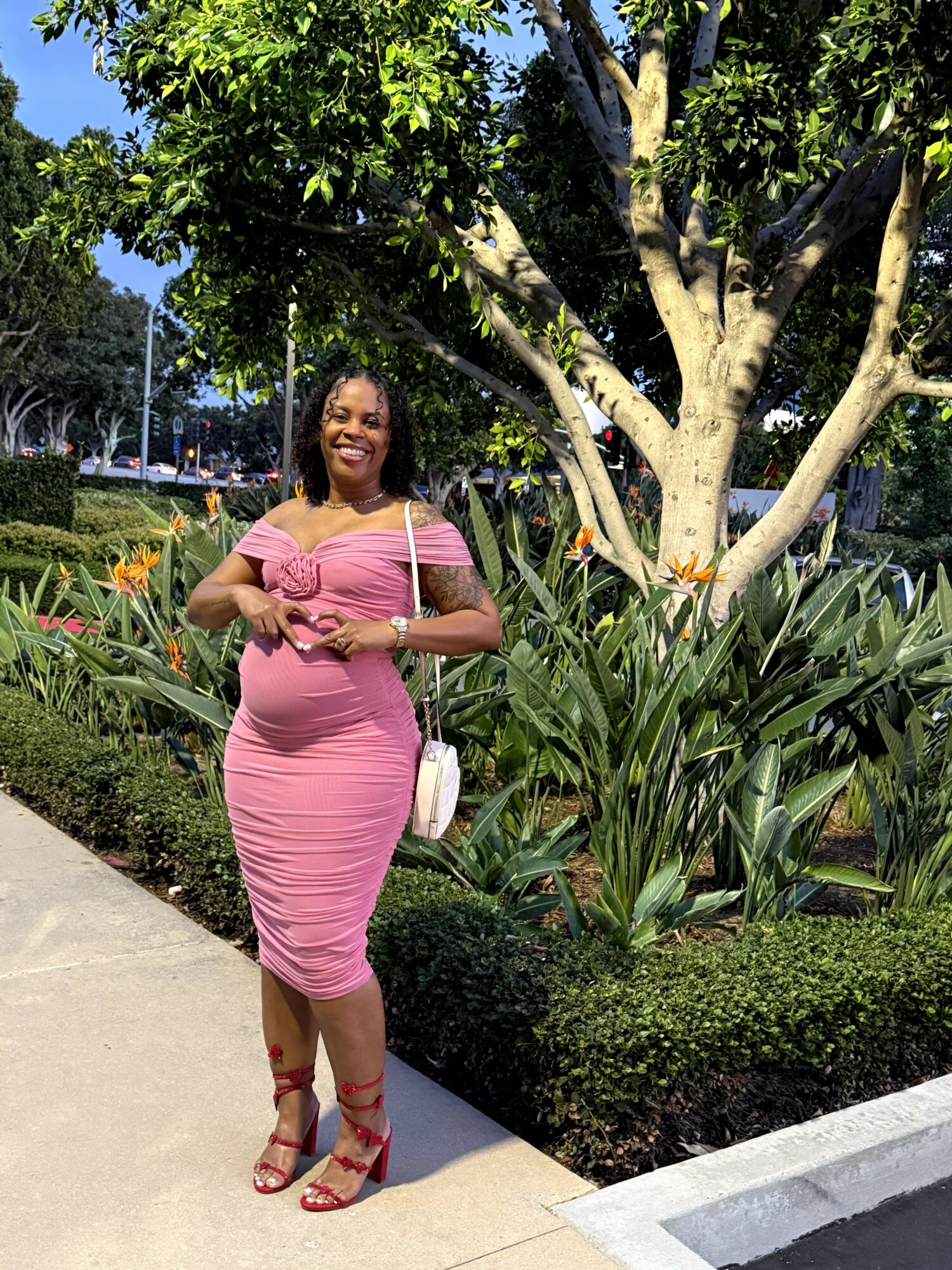 Smiling woman in pink dress standing on sidewalk with greenery and trees behind her.