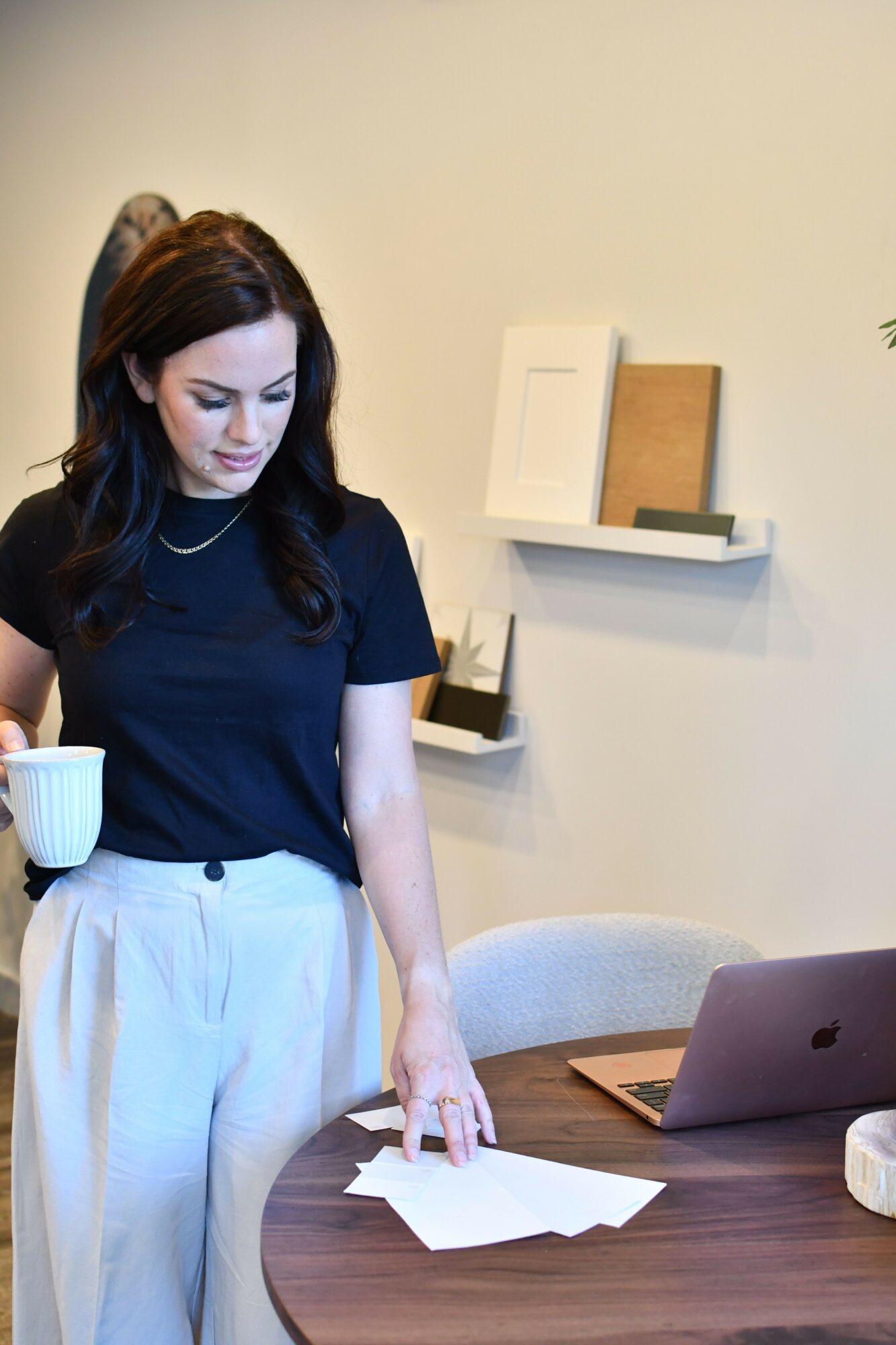 Woman with dark hair in a navy shirt and white pants stands near a table with papers and a laptop.