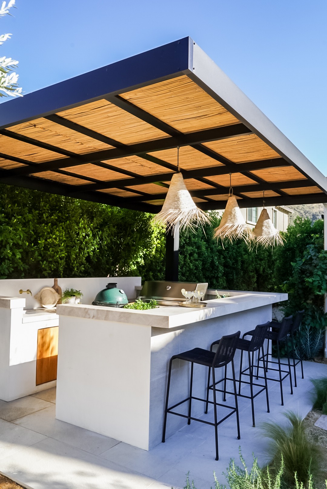 Outdoor kitchen with white counter, black bar stools, hanging pendant lights, and green plants in background.