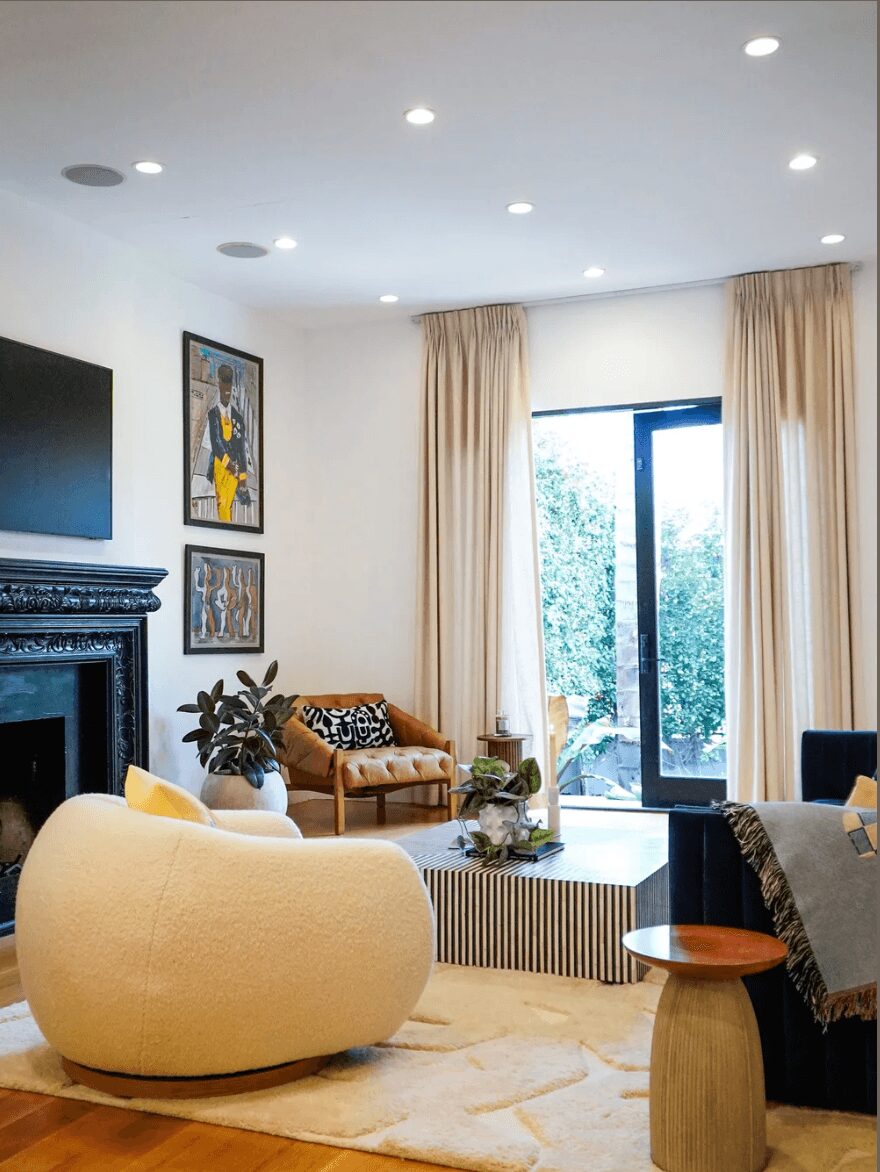 Living room with beige curtains, a large window, a beige armchair, a wooden table, and artwork on the wall.