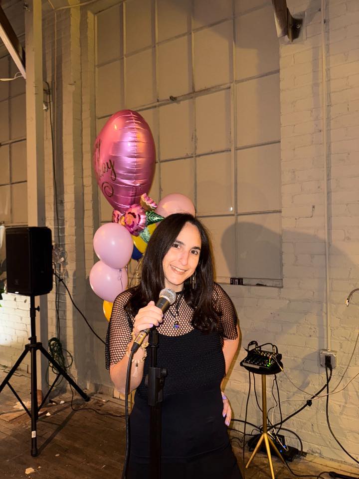 Young woman with dark hair holding a microphone, smiling, standing indoors near balloons and equipment.
