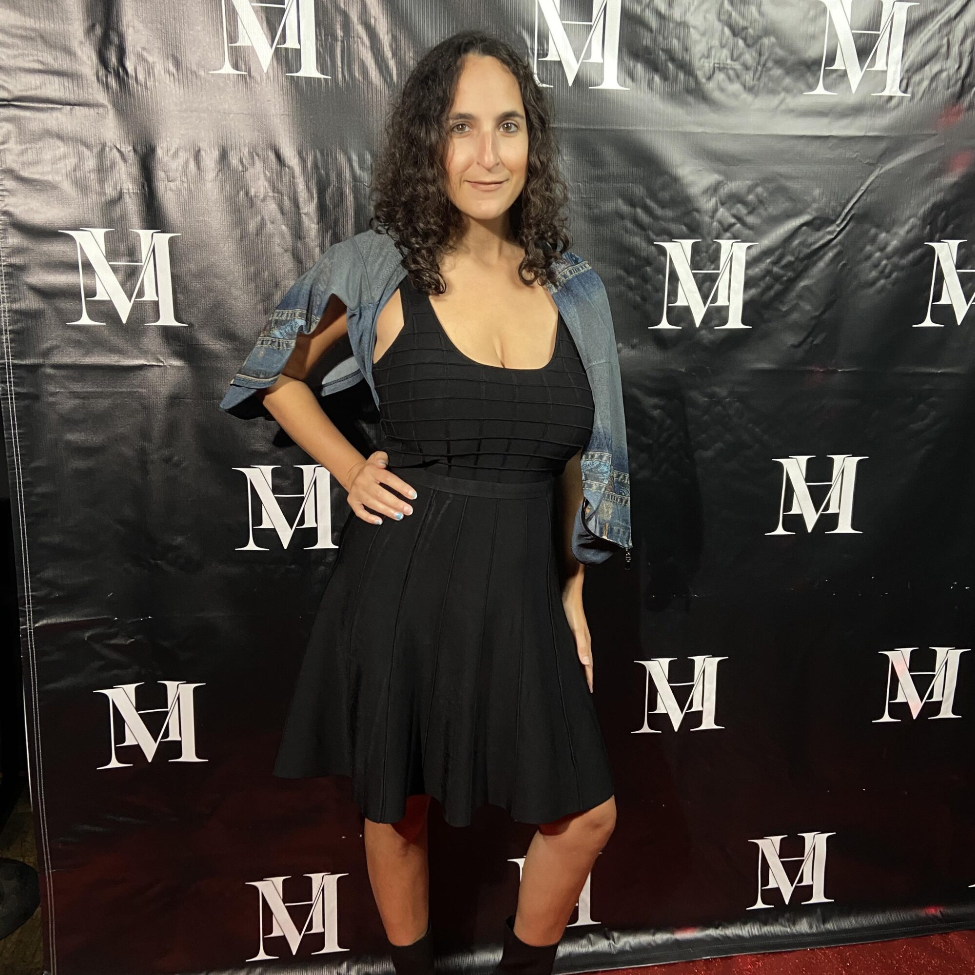 Woman with curly dark hair in black dress and denim jacket stands in front of black backdrop with white 'M' logos.