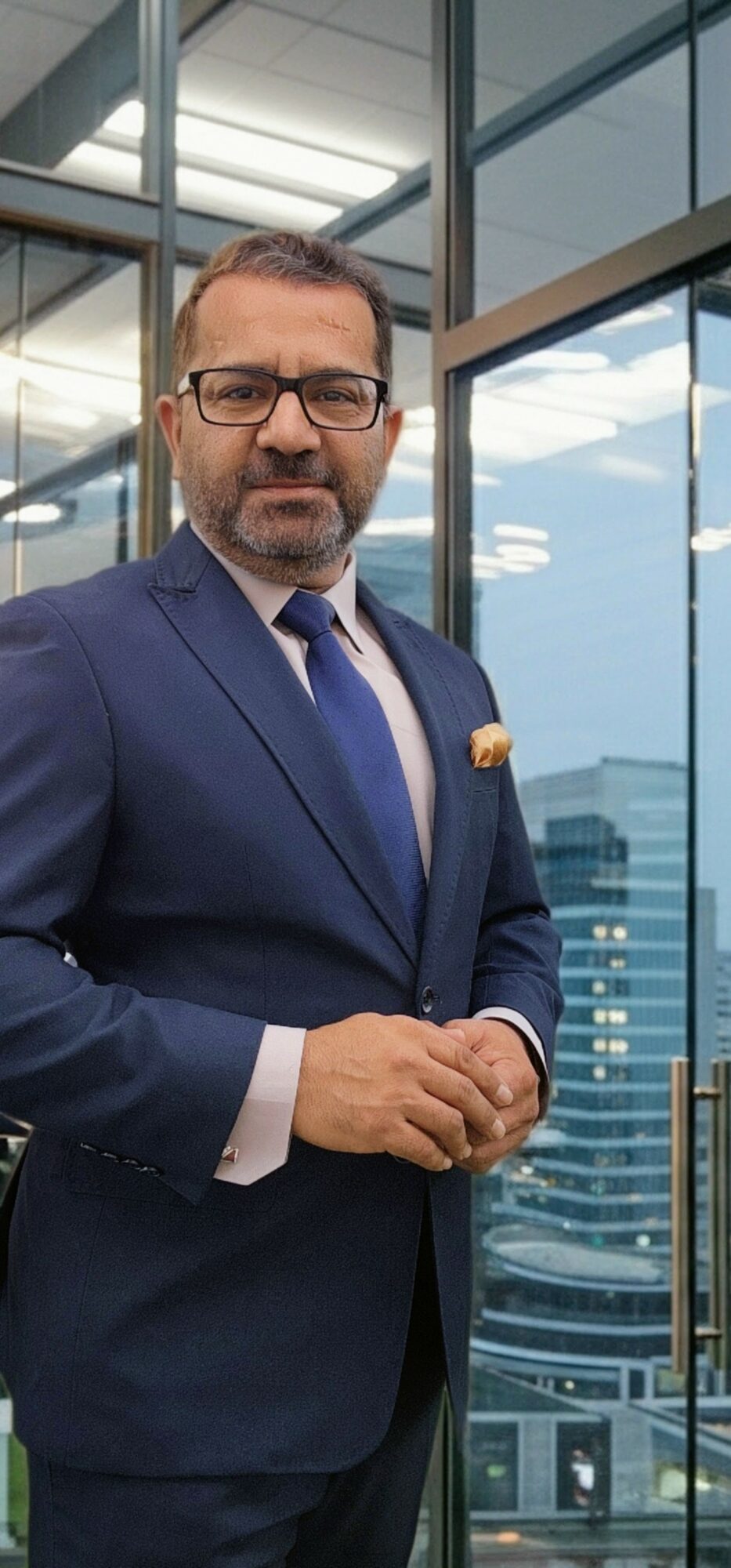 Man in suit and glasses standing in modern office building with glass windows and cityscape background.