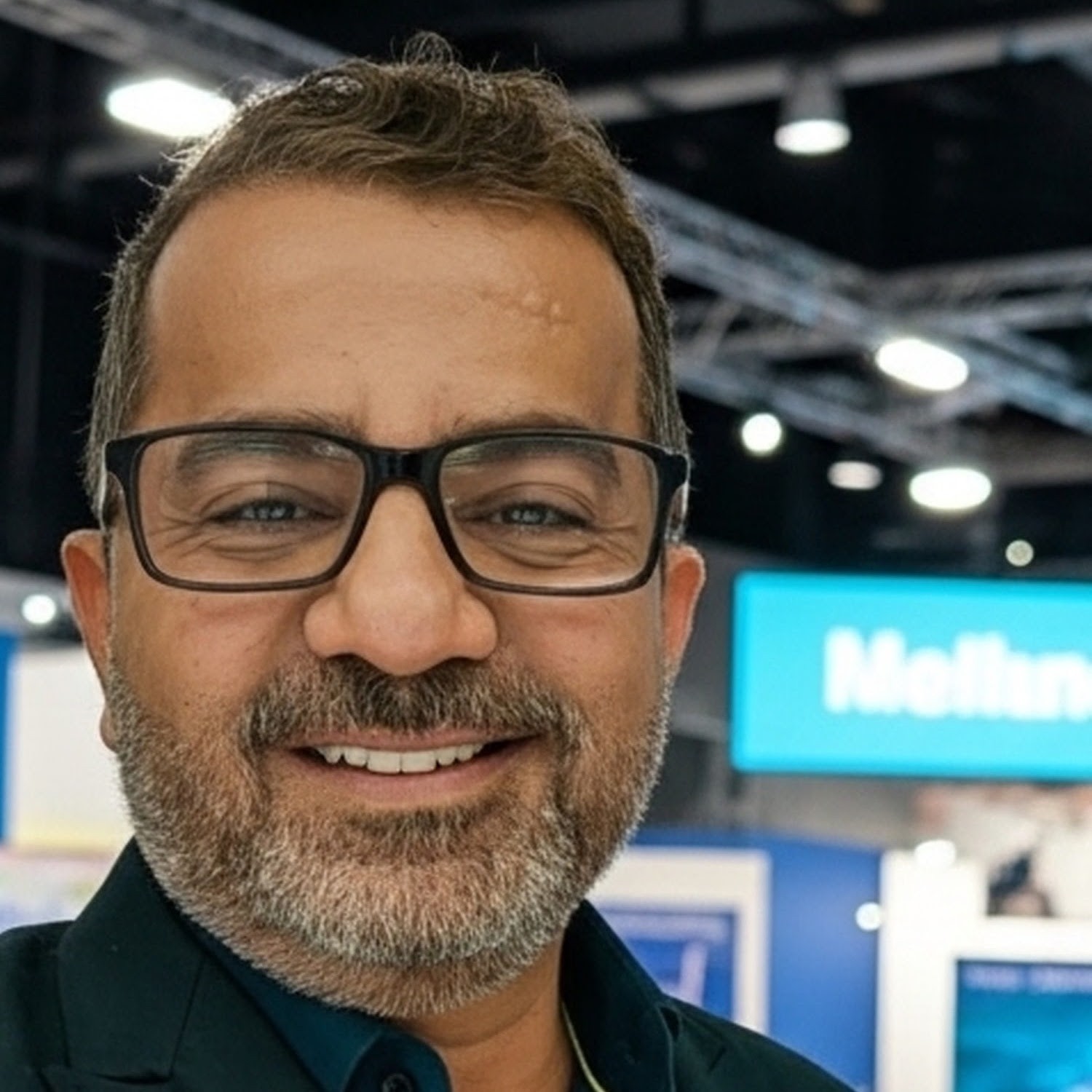 Man with glasses smiling, indoor setting with bright lights and a blue sign in the background.
