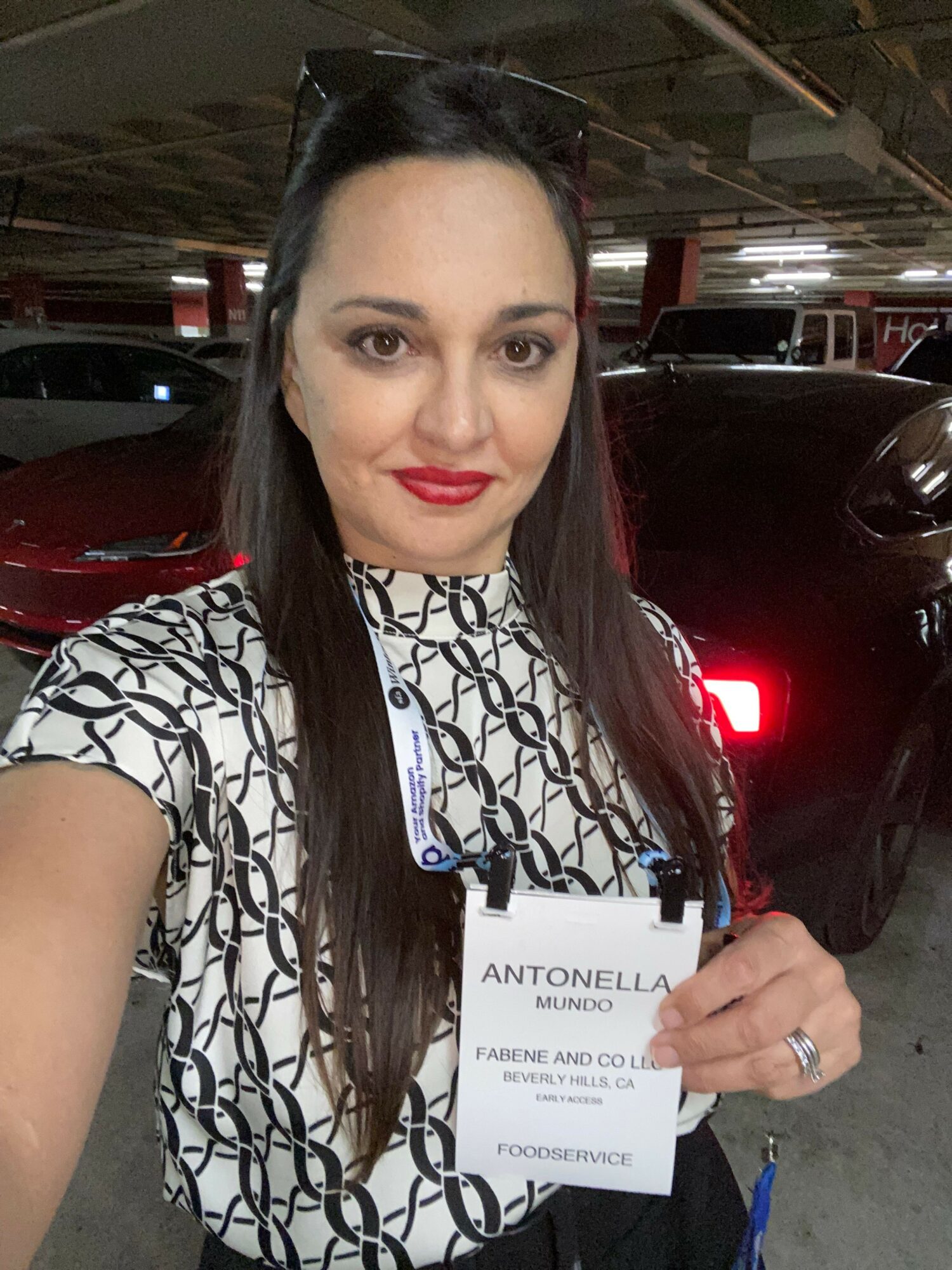 Woman with long dark hair, red lipstick, wearing a patterned top, holding a badge, in parking garage.