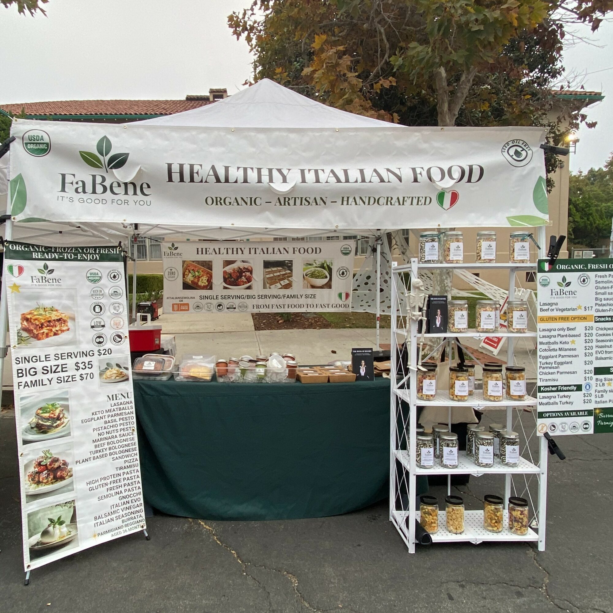 Market stall with a white canopy displaying 'Healthy Italian Food' and various food items on shelves and tables.