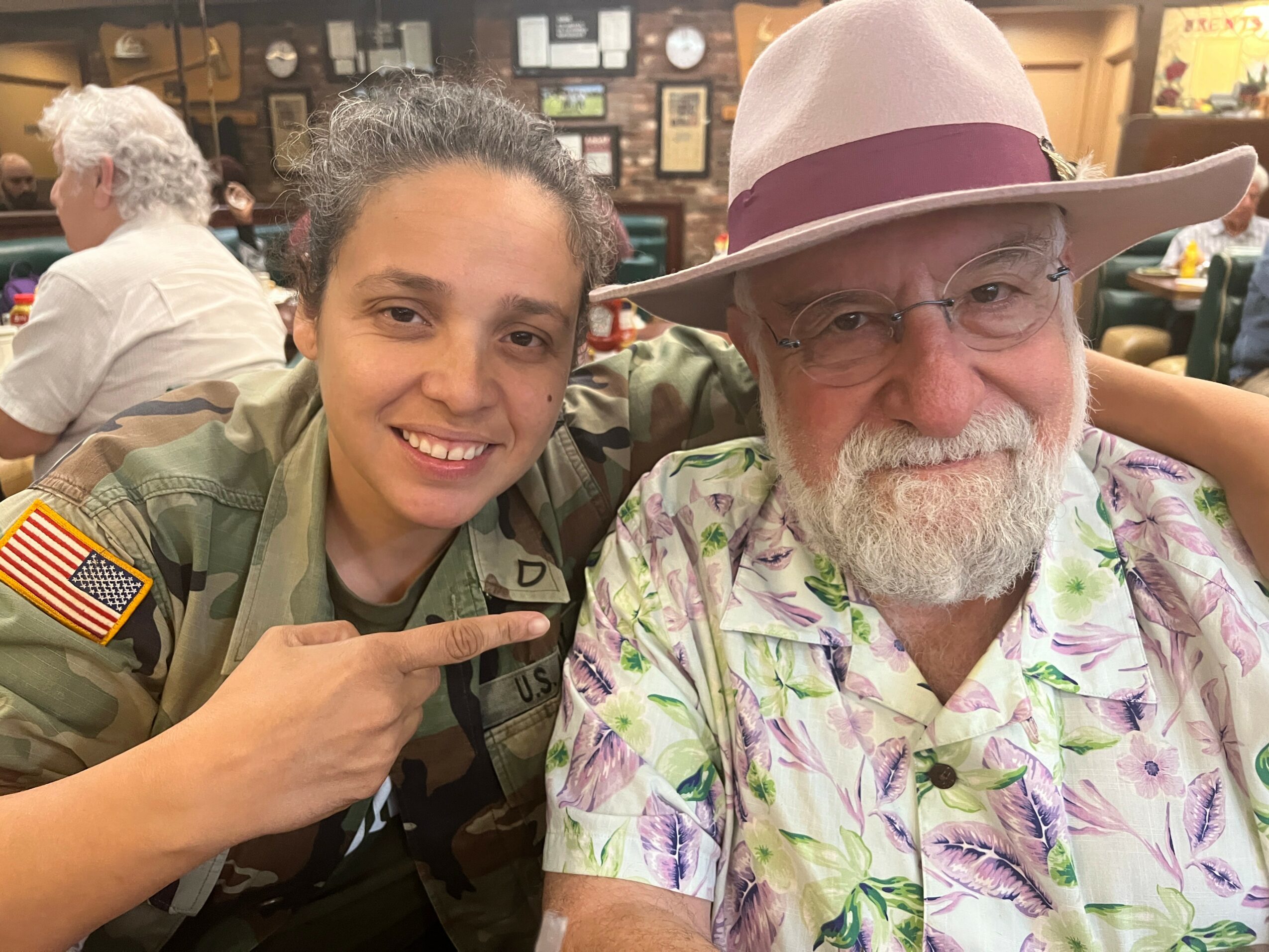 Young woman in military uniform smiling with older man in a pink hat and floral shirt, pointing at him.
