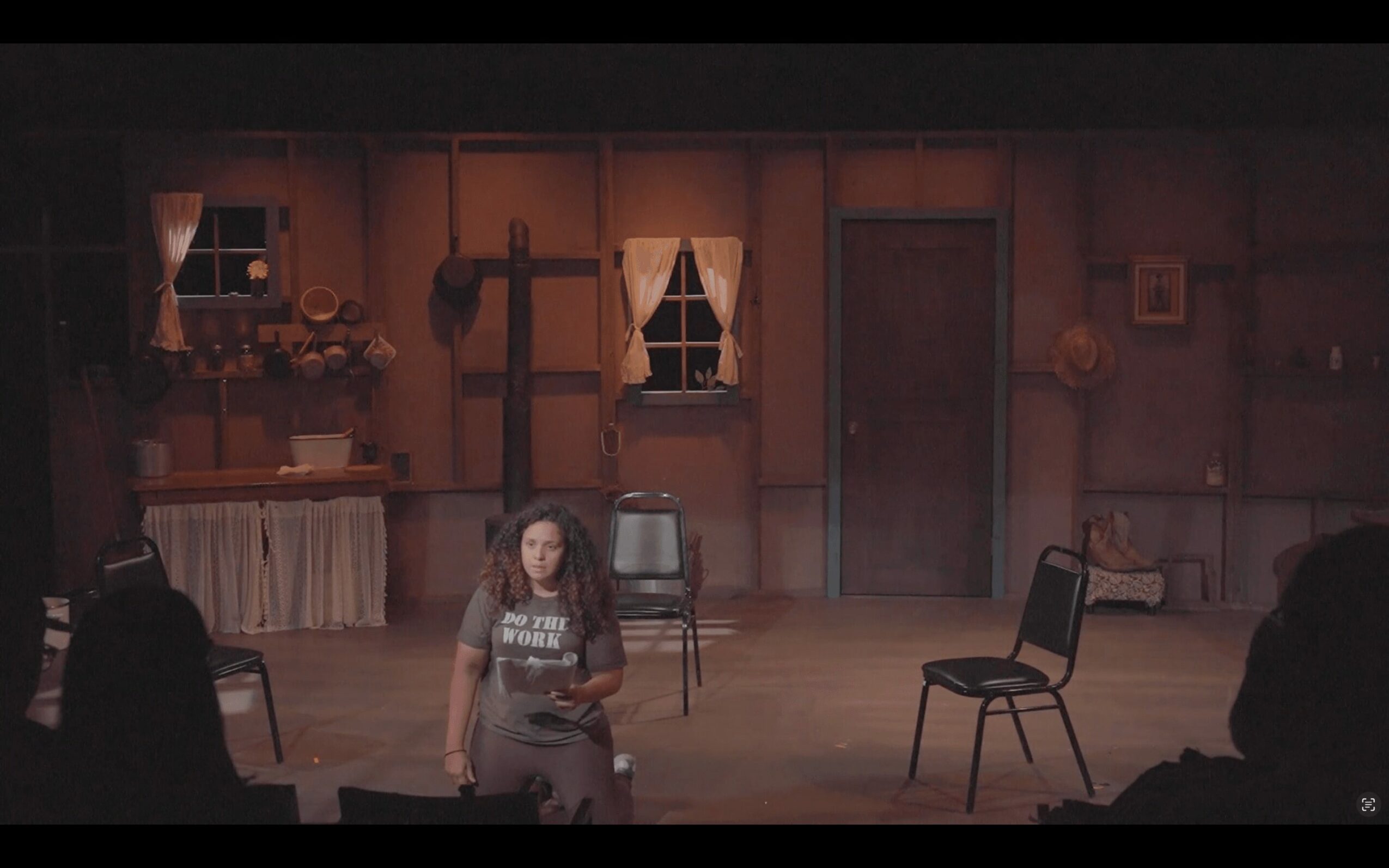 Stage scene with a woman sitting on the floor, three chairs, and a wooden backdrop with windows and decorations.