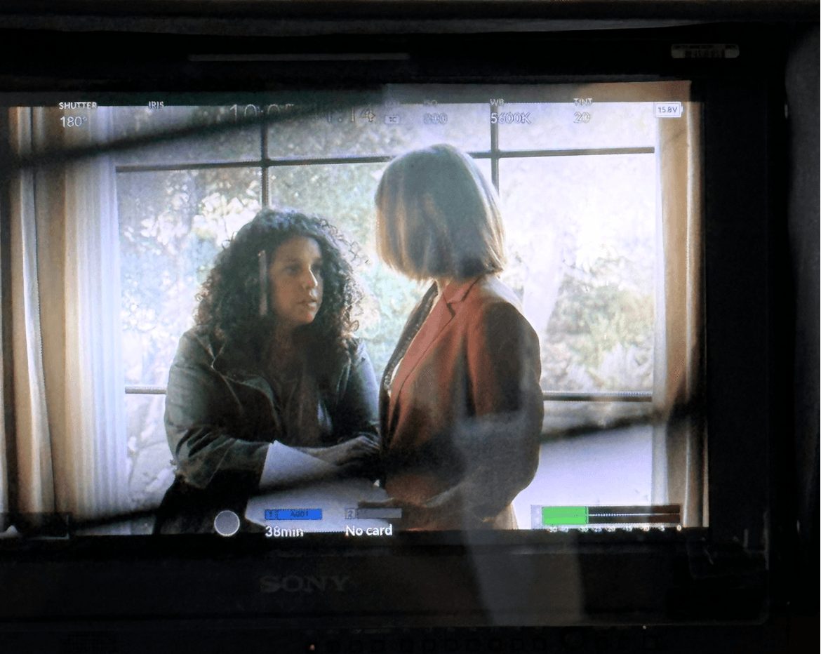 Two women face each other in front of a window with trees outside, engaged in conversation.