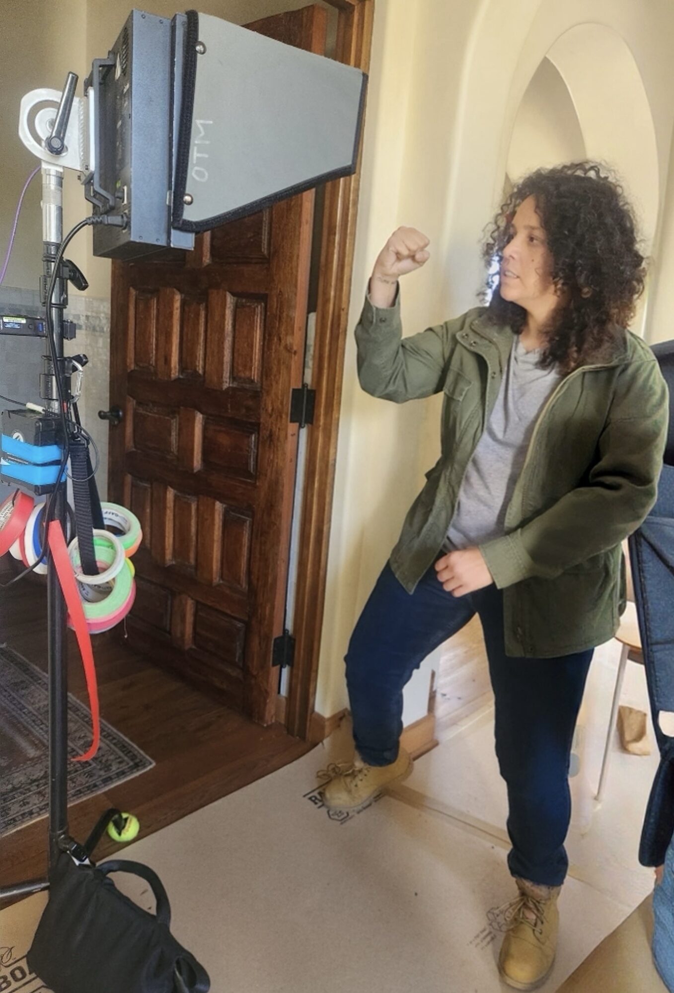 Woman with curly hair standing indoors, gesturing with her right hand, near a door and a large camera or monitor.