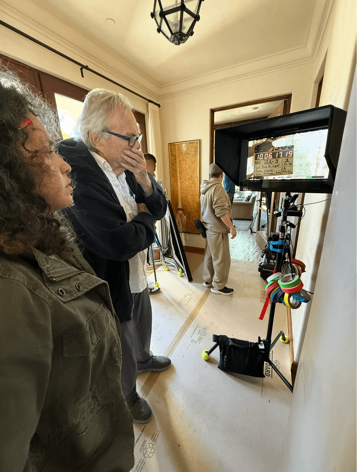 Three people observing a monitor on a stand in a room with cream-colored walls and a chandelier.