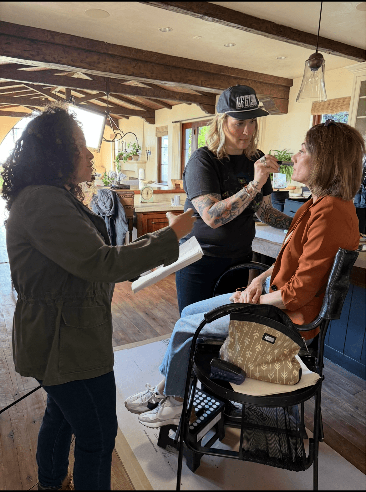 Makeup artist applies makeup to a woman seated in a chair, with a person holding a notepad nearby in a cozy room.