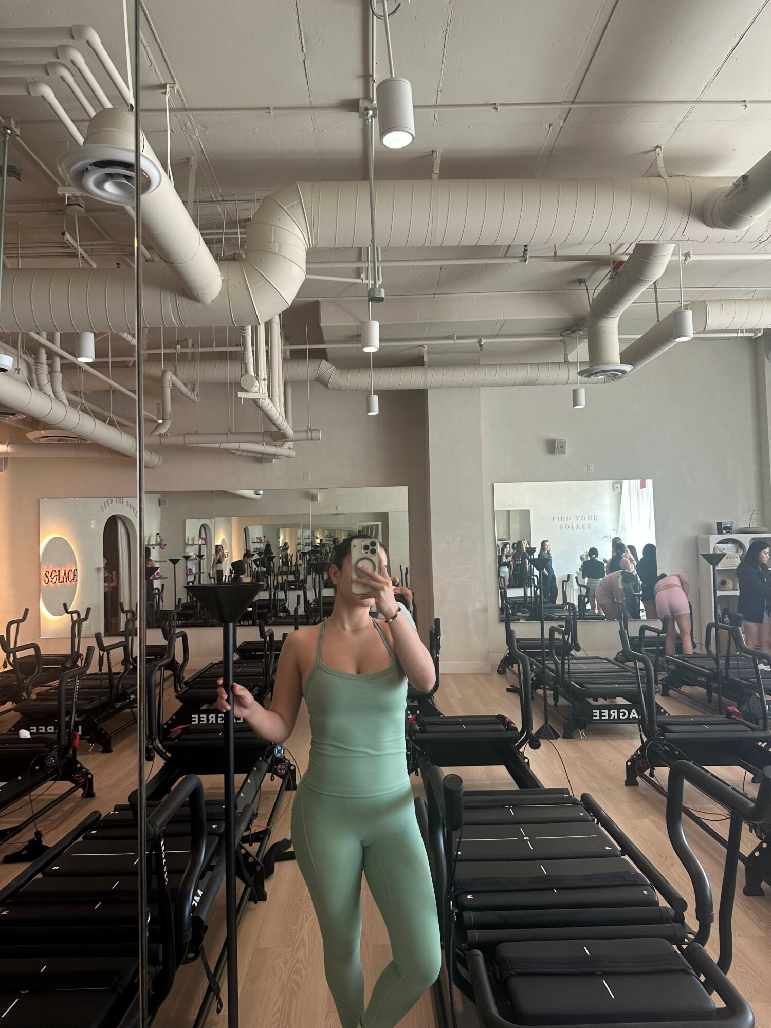 Person in light green workout outfit taking a mirror selfie in a gym with exercise equipment and other people.