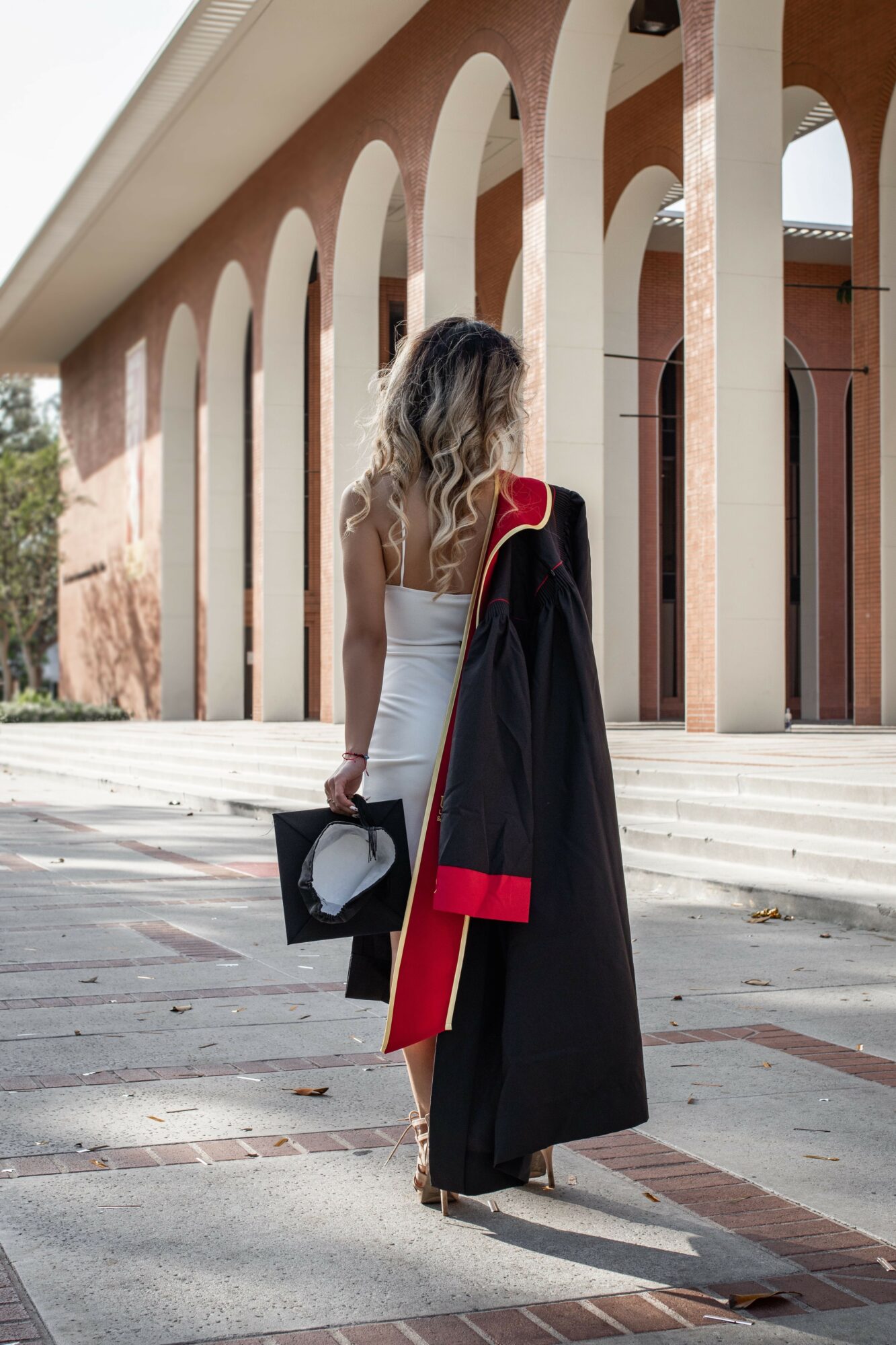 Woman in graduation gown holding a cap and diploma, standing outside near a building with arches.