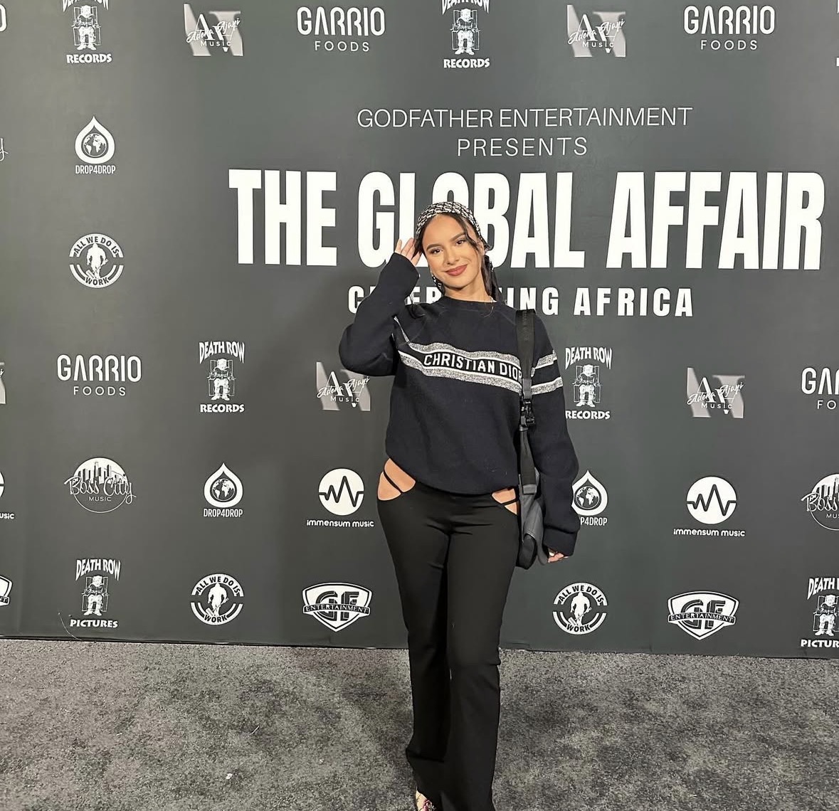 Woman standing in front of a backdrop with event title and logos, smiling and posing with one hand on her head.