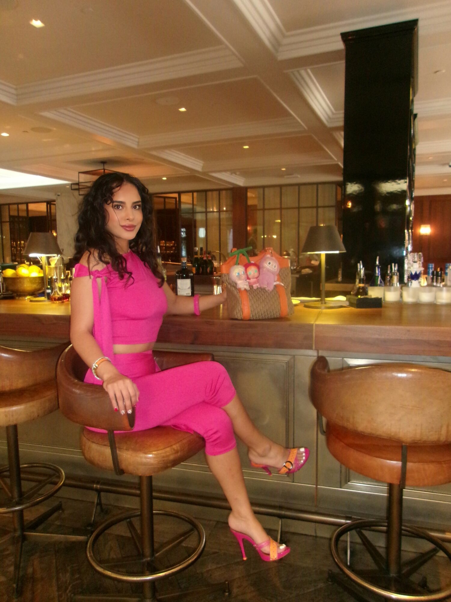Woman in pink outfit sitting on bar stool in a restaurant or bar setting with a cake in the background.