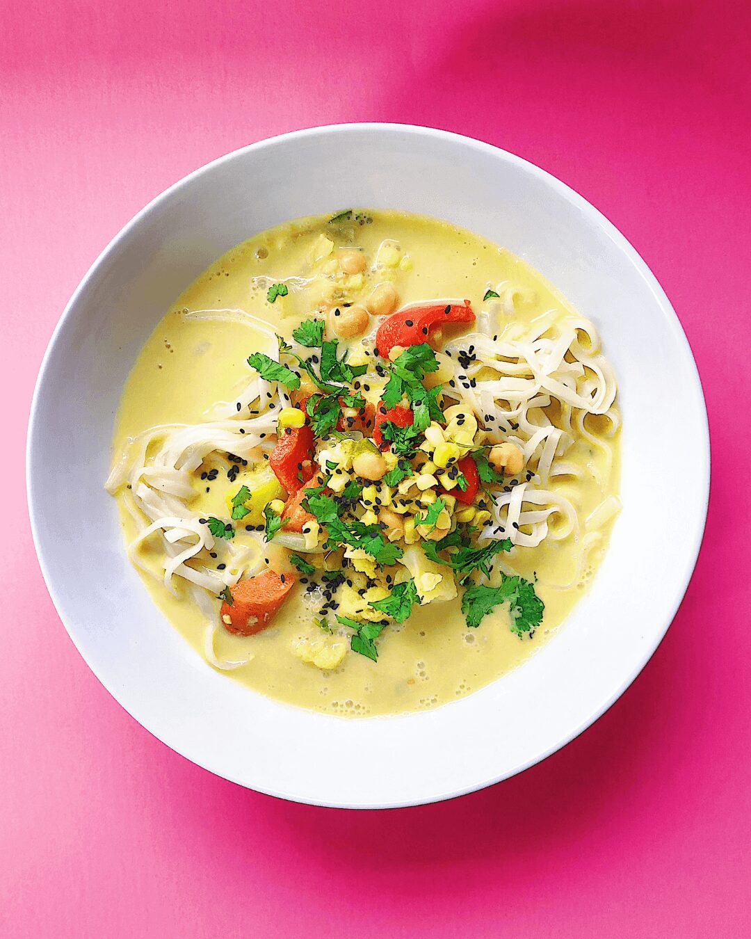 Bowl of creamy soup with noodles, cherry tomatoes, herbs, and corn on a pink background.