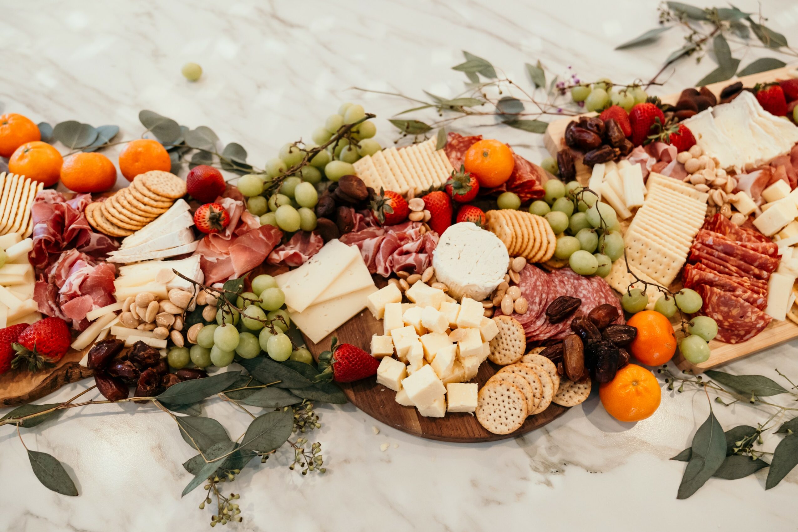Arrangement of various cheeses, fruits, and crackers on a white surface with greenery.