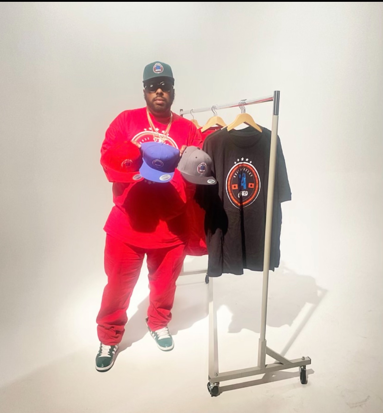 Person wearing red outfit holding three hats, standing next to a clothing rack with a black t-shirt, against a plain white background.