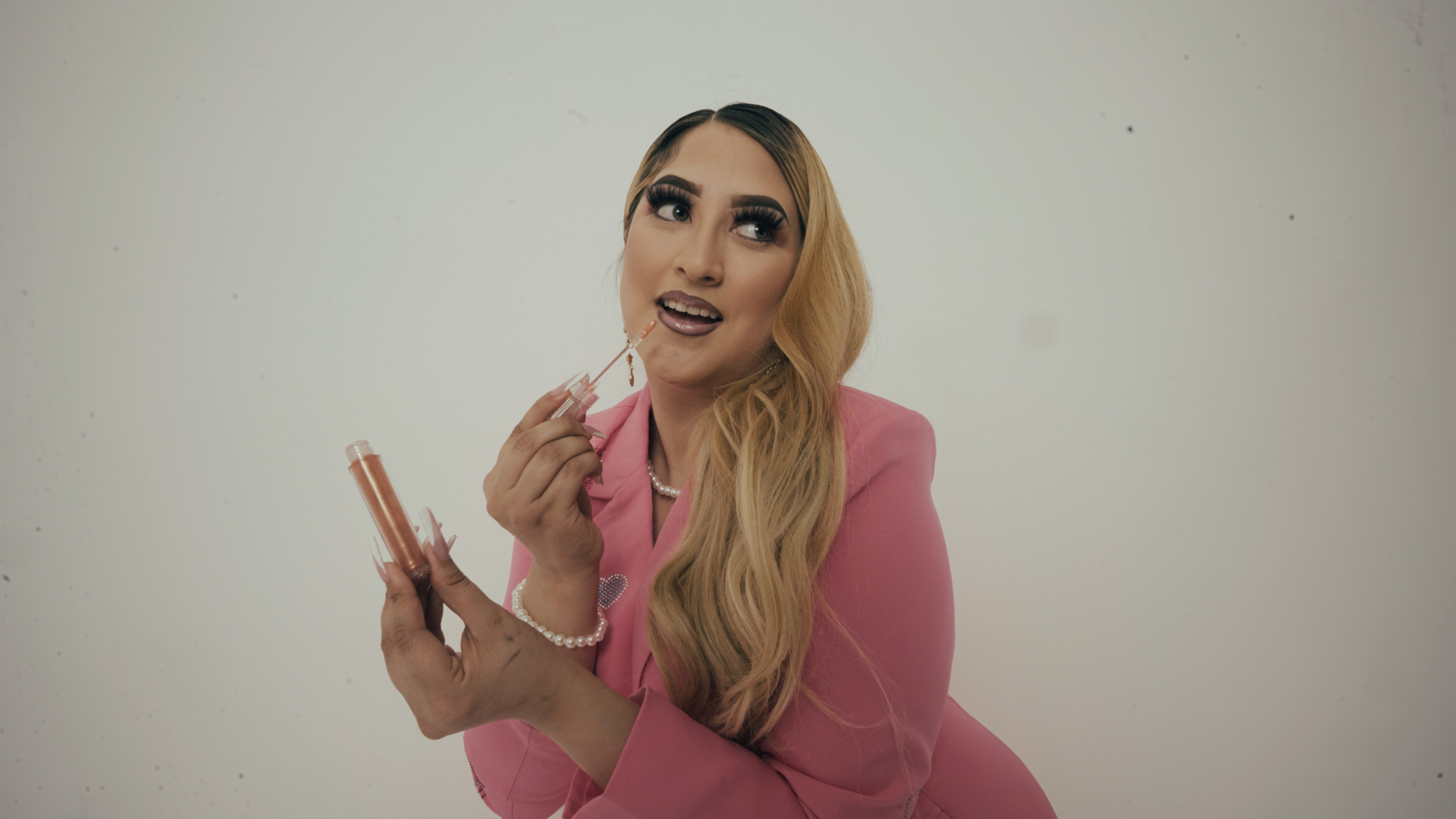 Woman with long blonde hair wearing pink blazer holding makeup items and smiling.