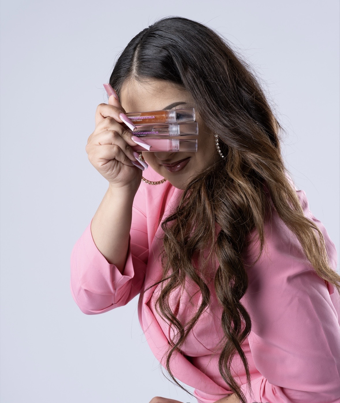 Woman with long wavy hair wearing pink top, holding glasses to her face, smiling.