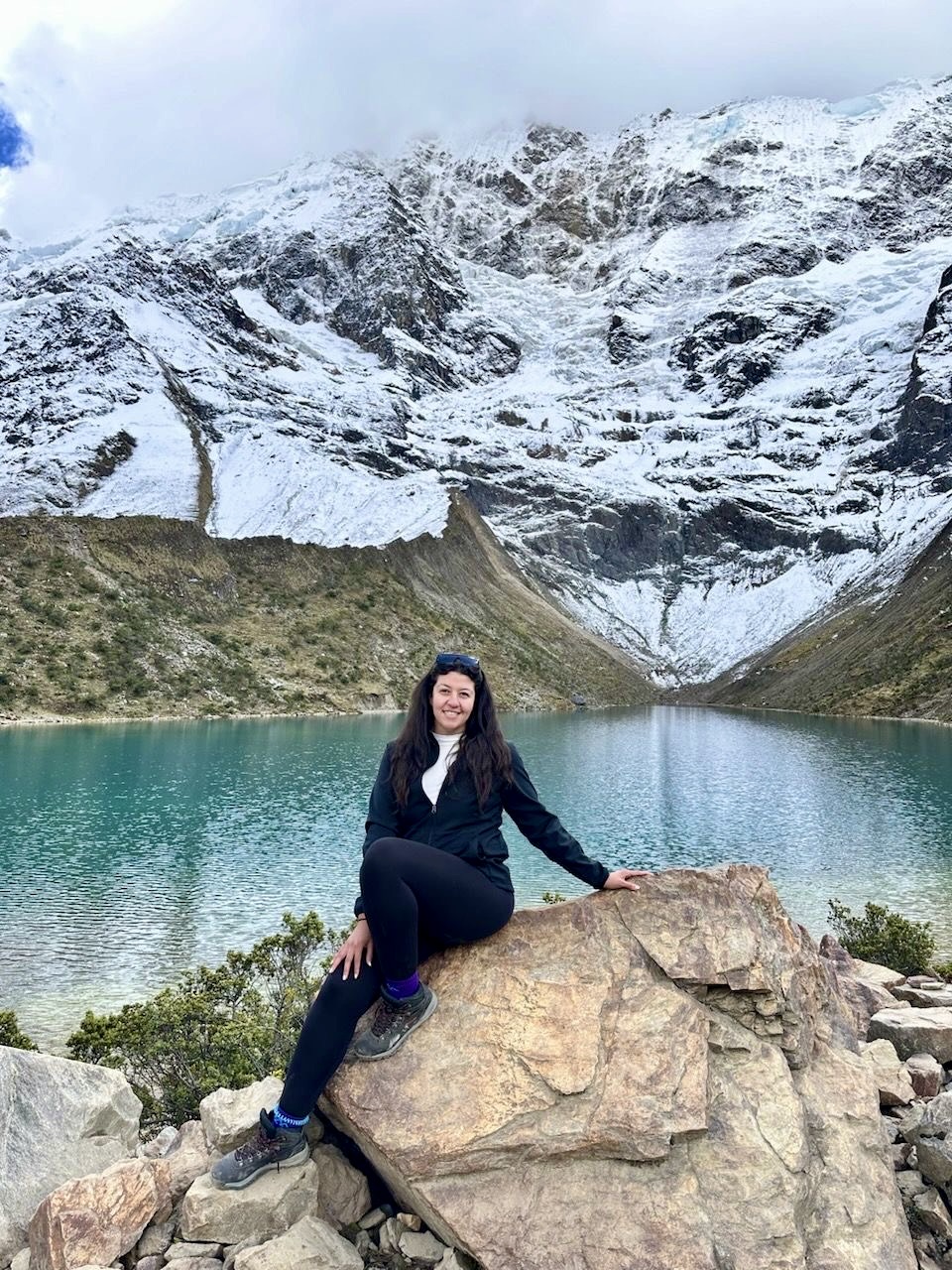 Woman sitting on a large rock by a turquoise lake with snow-covered mountains in the background.