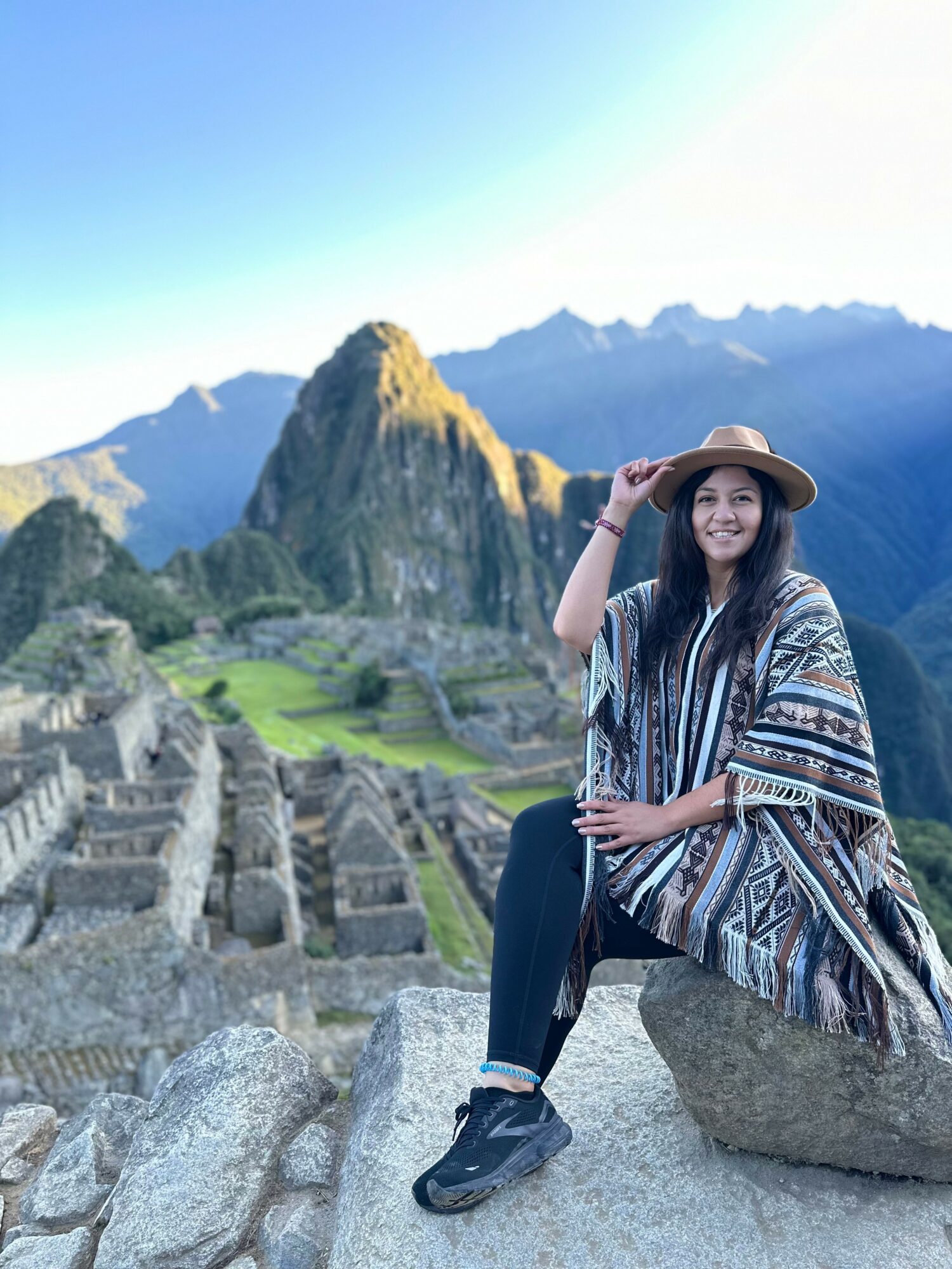 Woman sitting on a rock at Machu Picchu with mountains and ruins in background, smiling and wearing a hat and patterned poncho.