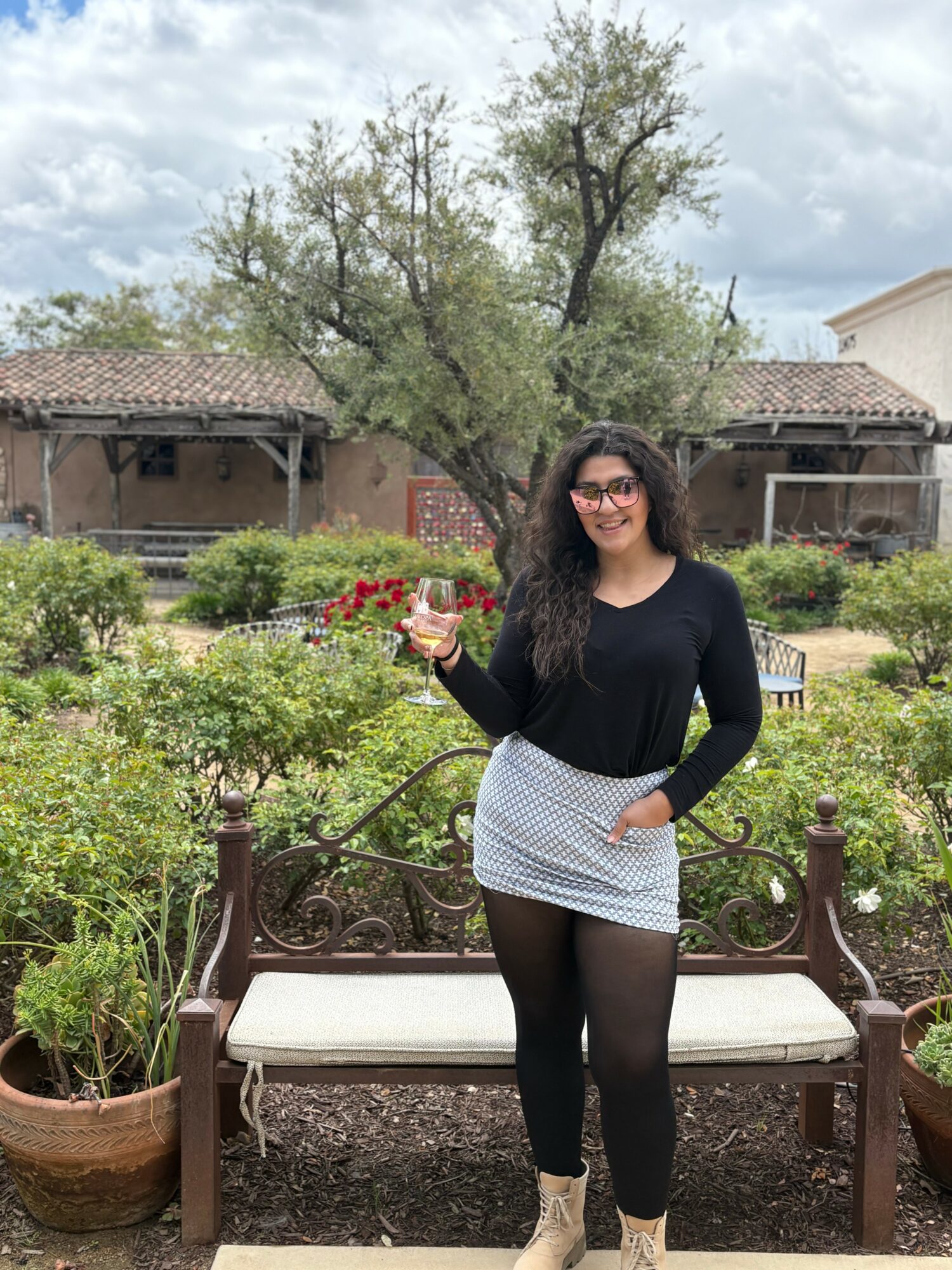 Woman with glasses holding a glass of wine, standing outdoors in a garden with trees and plants.
