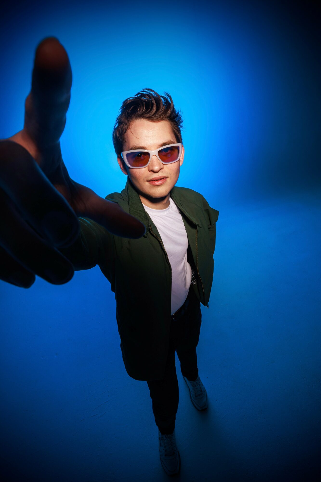 Person with sunglasses and green jacket reaching towards camera against blue background.
