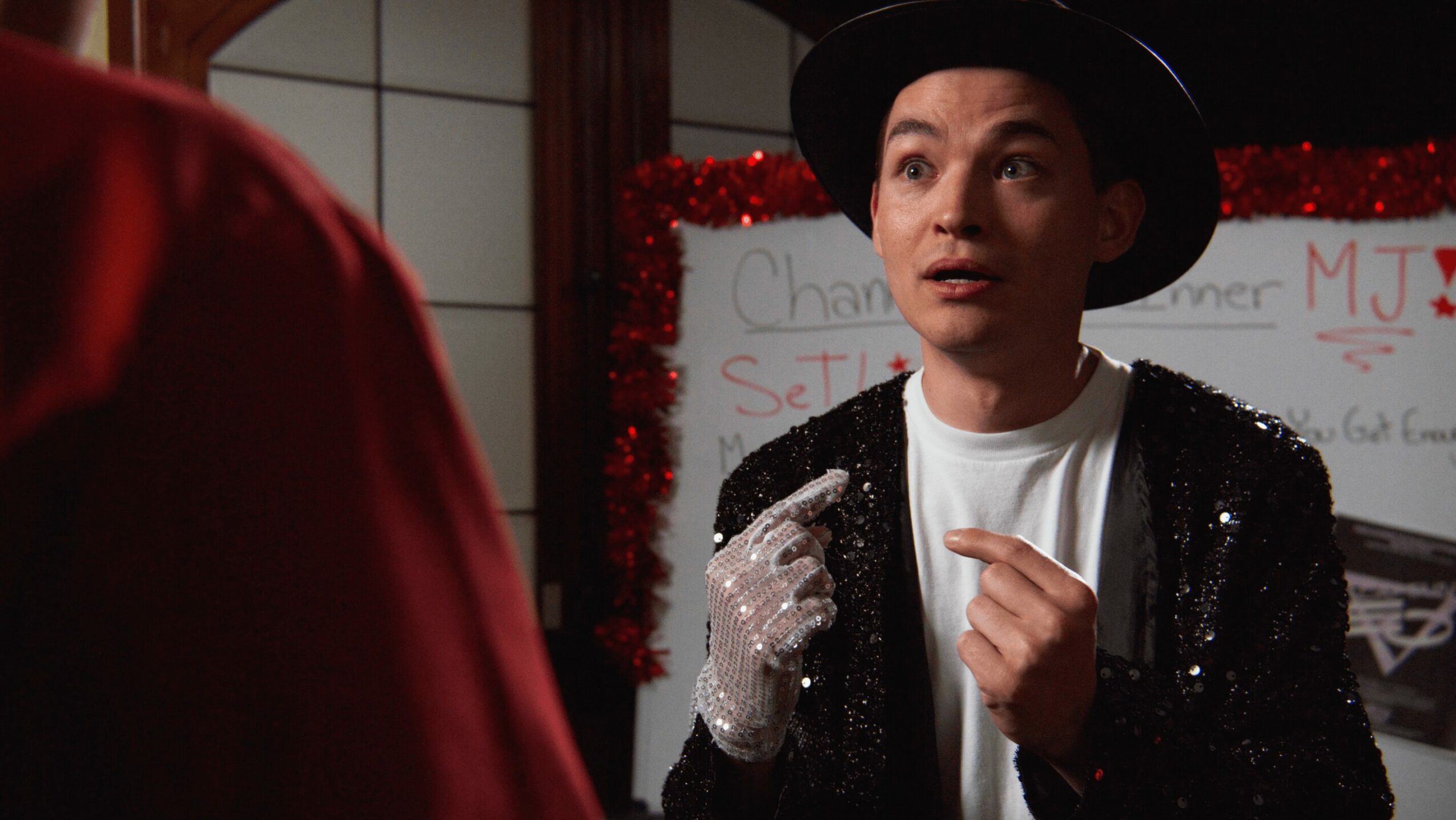 Person wearing a black hat and sequined jacket speaking, with a whiteboard and red decorations in the background.