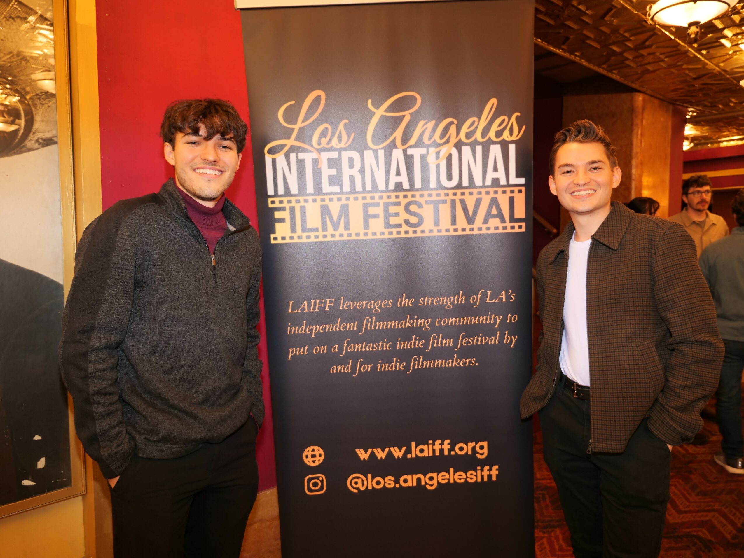 Two young men smiling and standing beside a Los Angeles International Film Festival banner indoors.