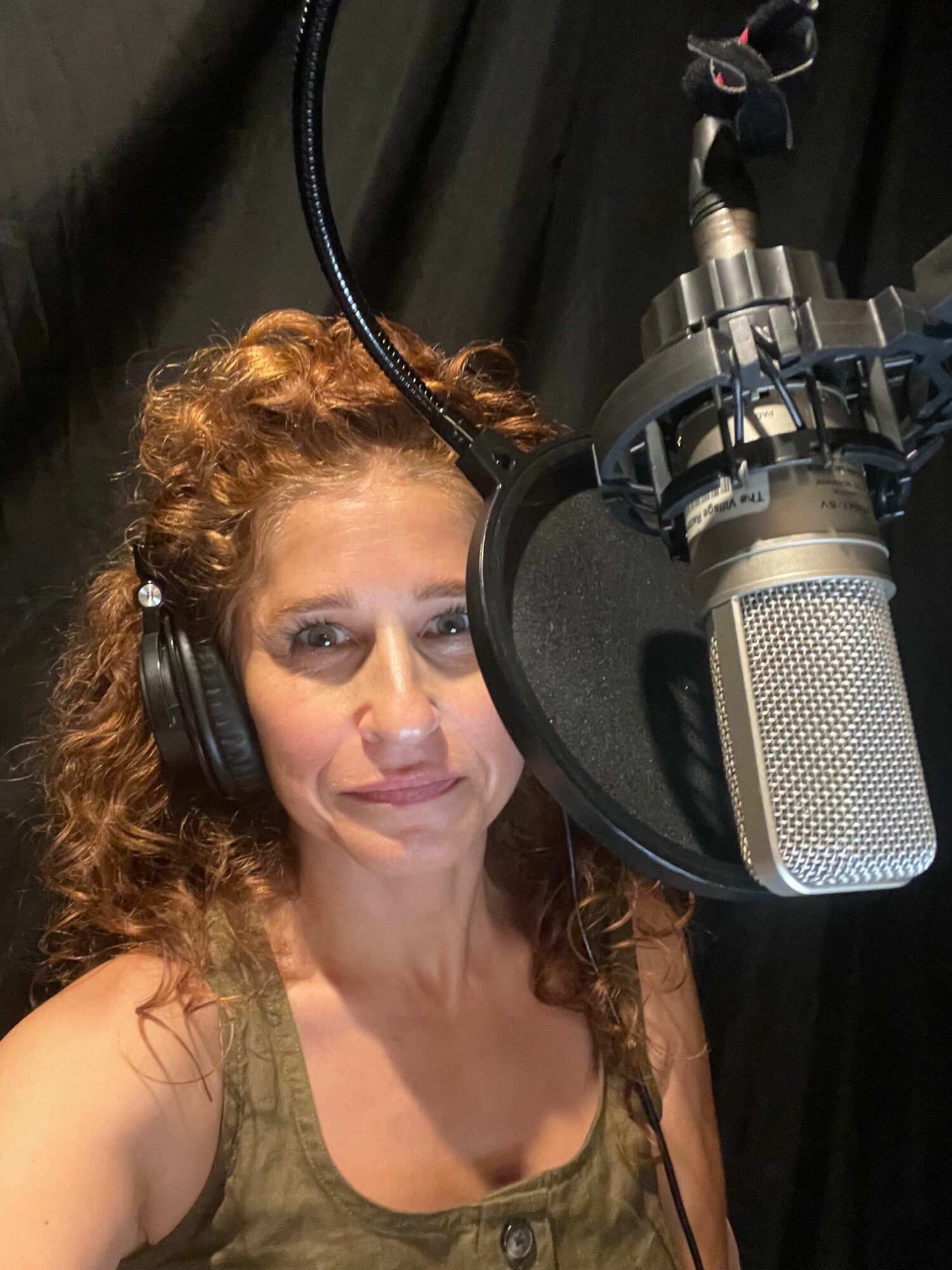 Woman with curly hair wearing headphones in recording studio with microphone.