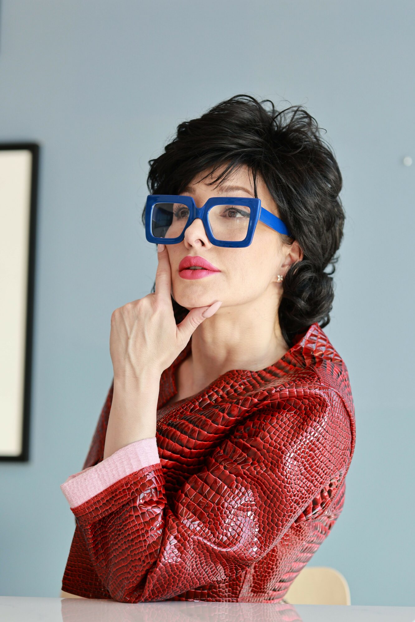 Woman with short dark hair and blue glasses, wearing a red textured jacket, touching her chin thoughtfully.