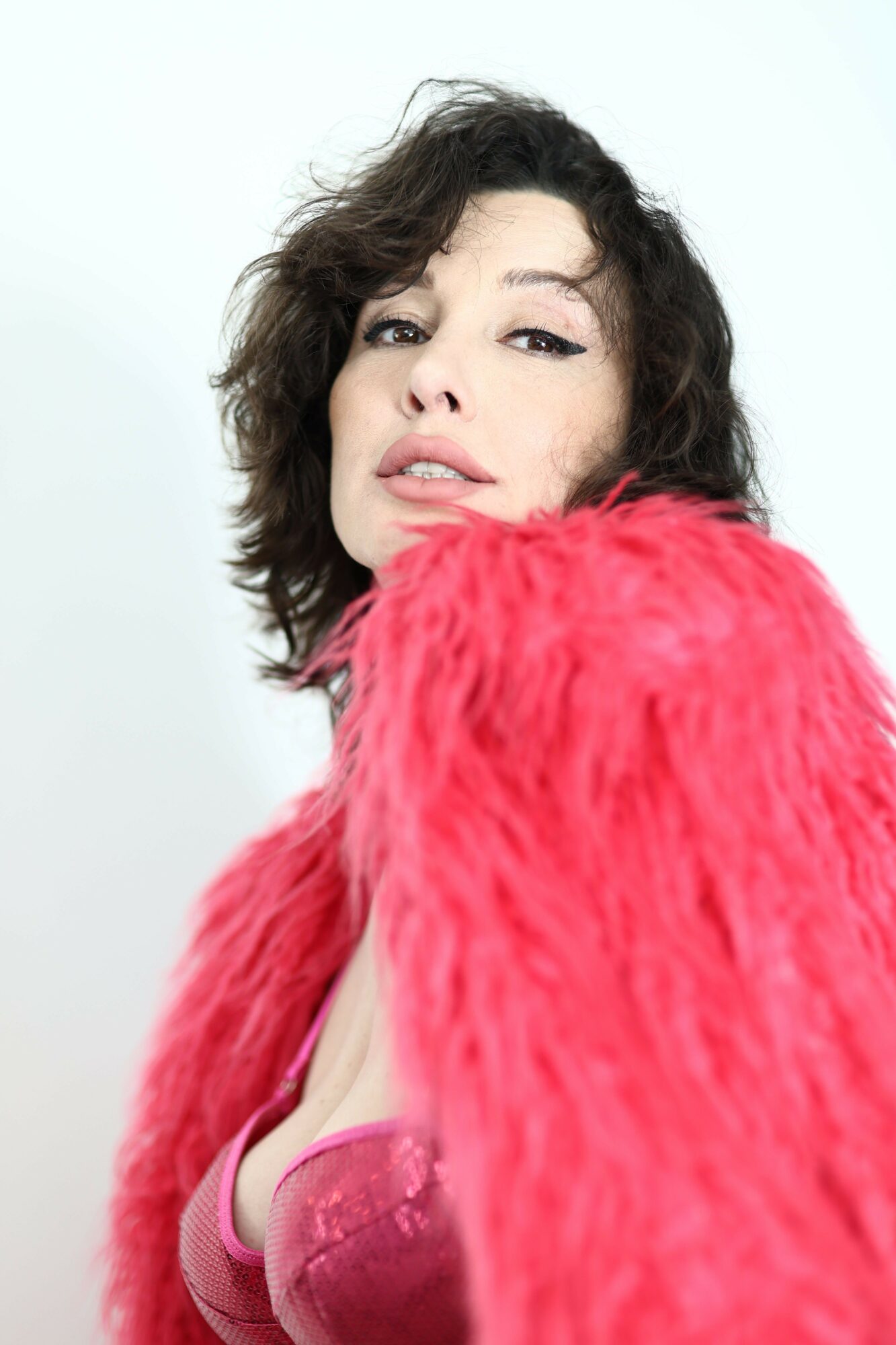 Woman with wavy hair wearing a pink fuzzy coat, looking at the camera against a plain white background.