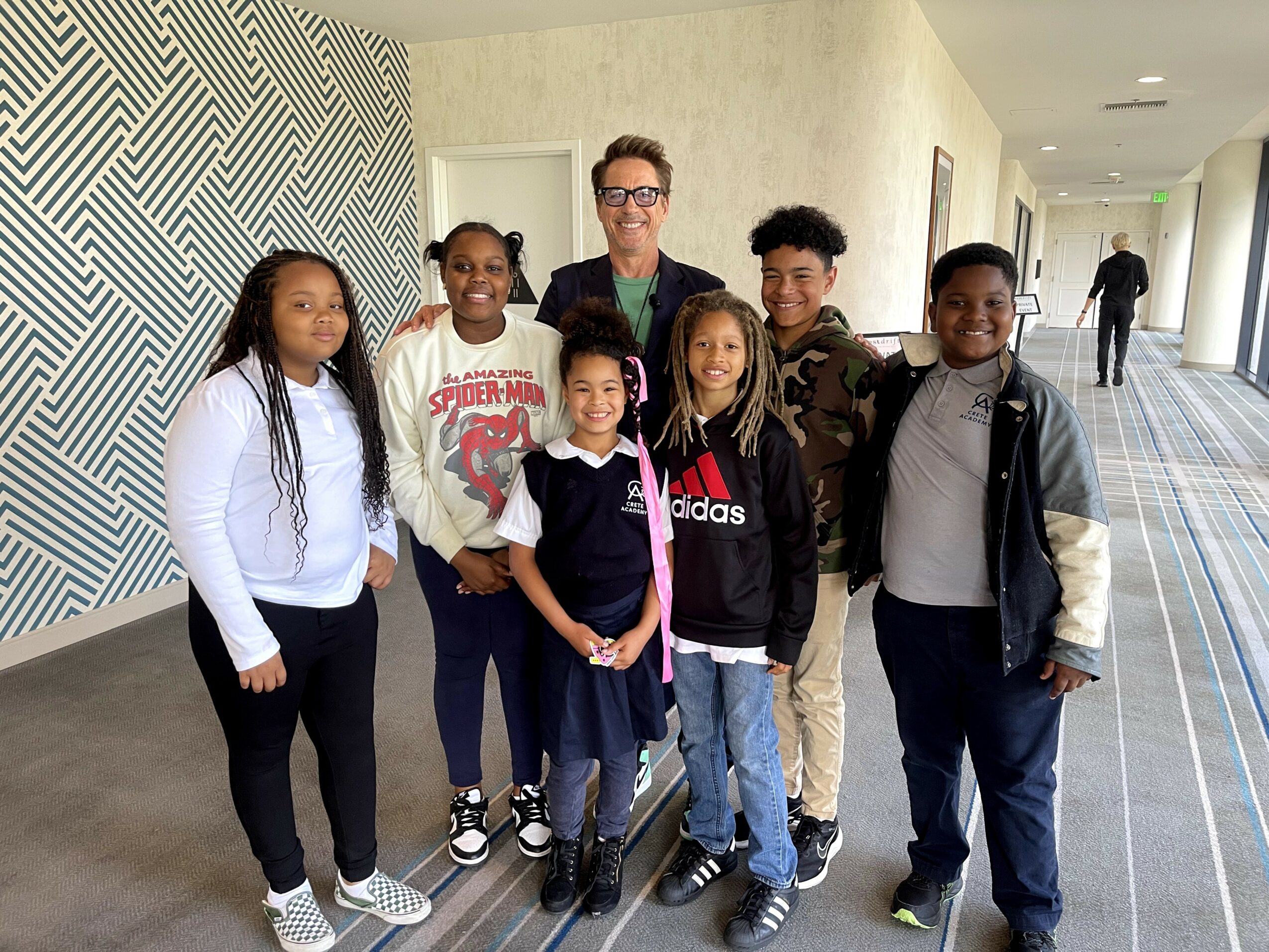 Group of seven children and one adult standing in a hallway, smiling for the photo.